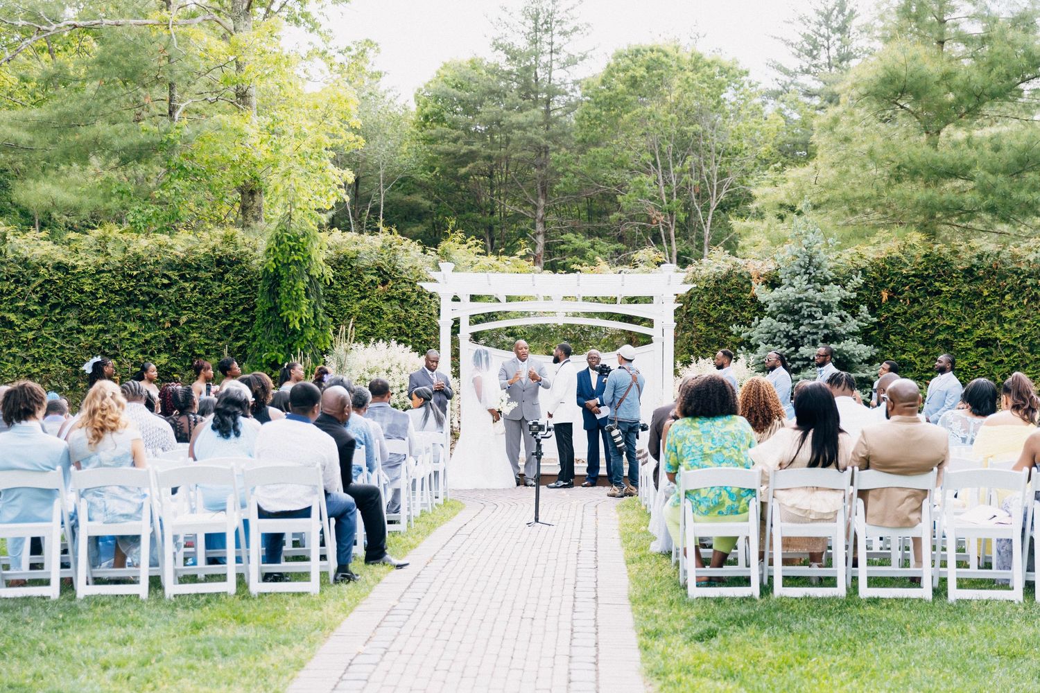 Outdoor garden wedding ceremony with guests seated in white chairs along a brick aisle leading to a white pergola altar.