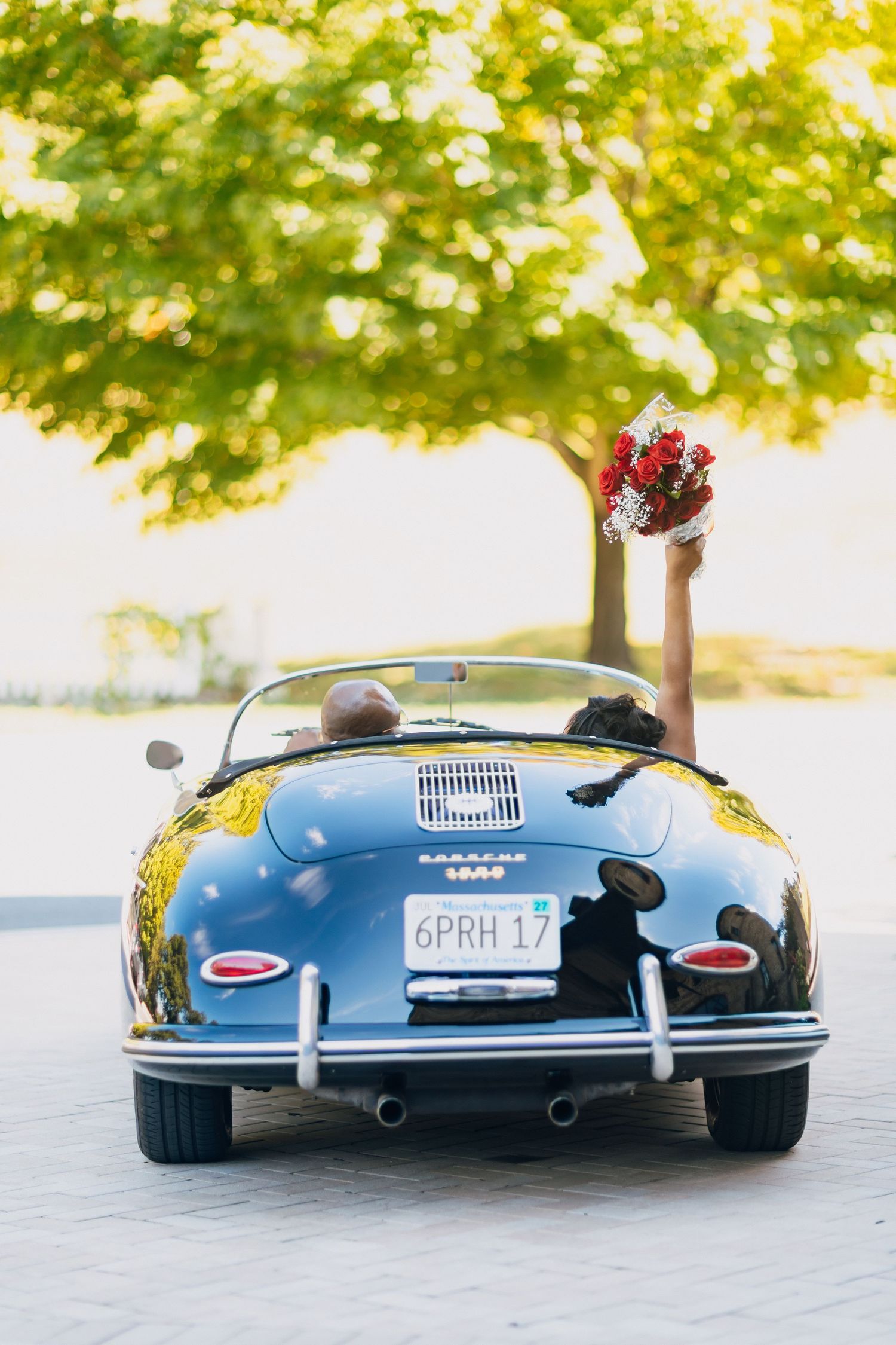 Newlyweds driving away in a classic black Porsche 356 convertible, bride raising red bouquet joyfully.