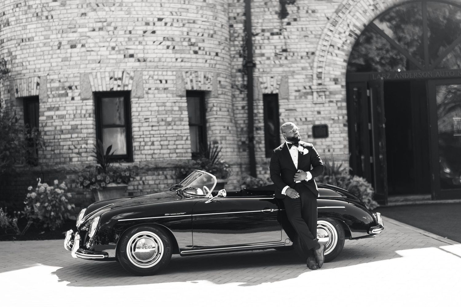 Dapper Black groom in tuxedo poses confidently beside a vintage black Porsche convertible in black and white.