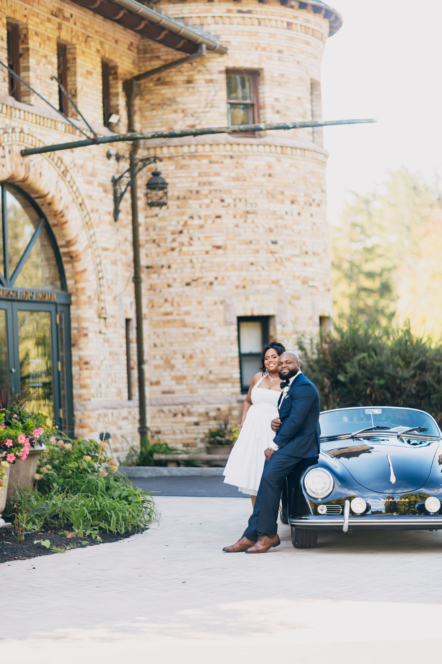 A newlywed couple embraces beside a vintage convertible in front of a historic stone tower.