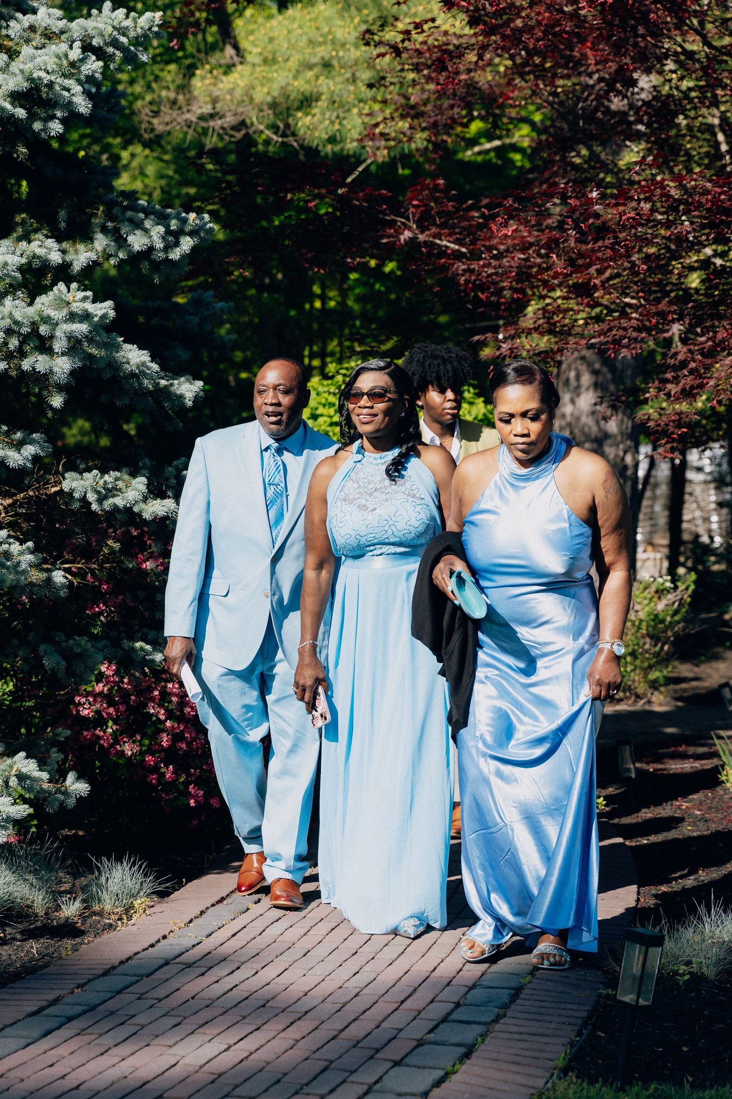 A group of four people in coordinating light blue formal attire walk along a garden path surrounded by lush greenery.