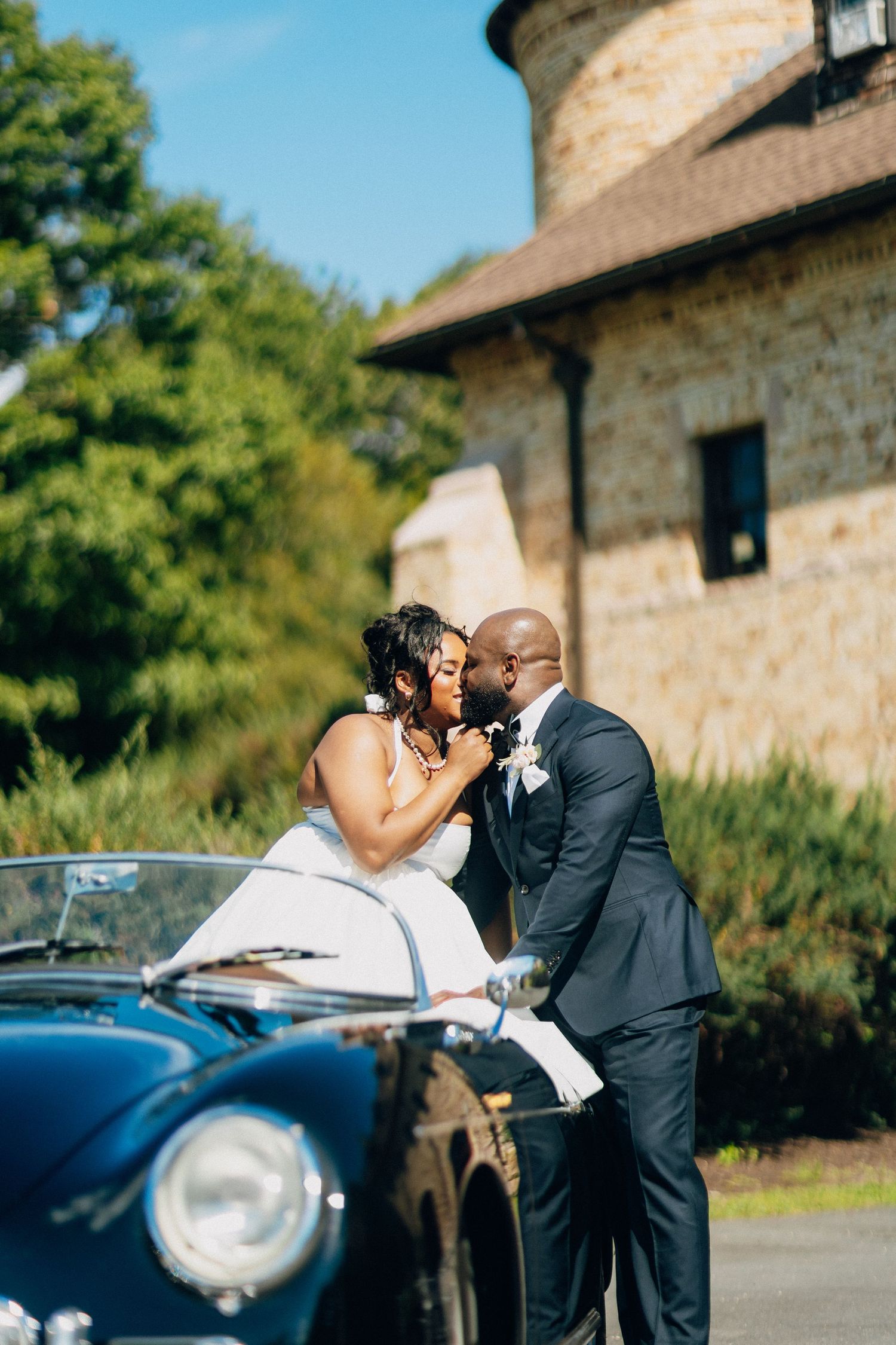 Bride and groom sharing a kiss in front of a classic black vintage Porsche at wedding venue.