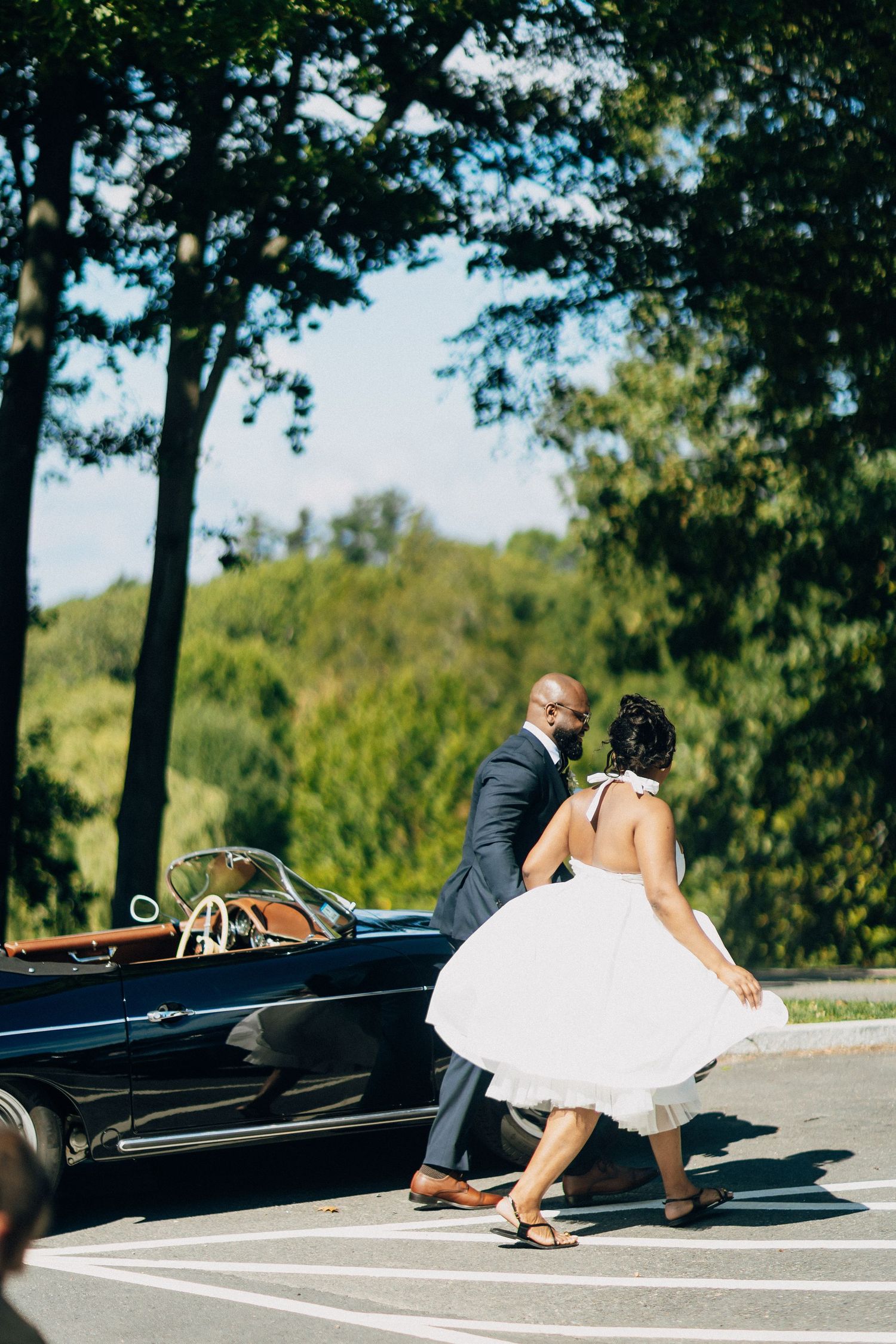 Bride and groom walking toward a vintage black convertible on a sunny day surrounded by lush green trees.