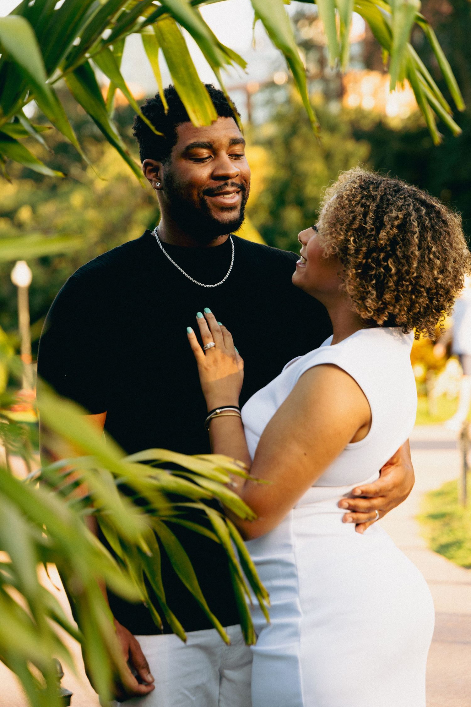 A joyful Black couple laugh together surrounded by lush tropical leaves in golden afternoon light.