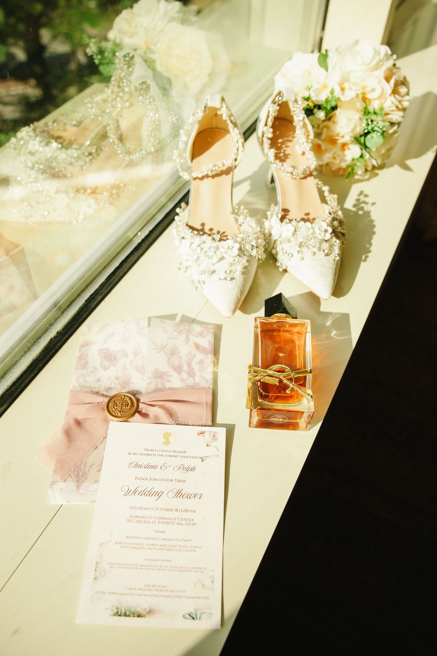 Bridal flat lay showcasing white floral lace heels, pink ribbon wedding invitation, perfume bottle, and white flower bouquet.