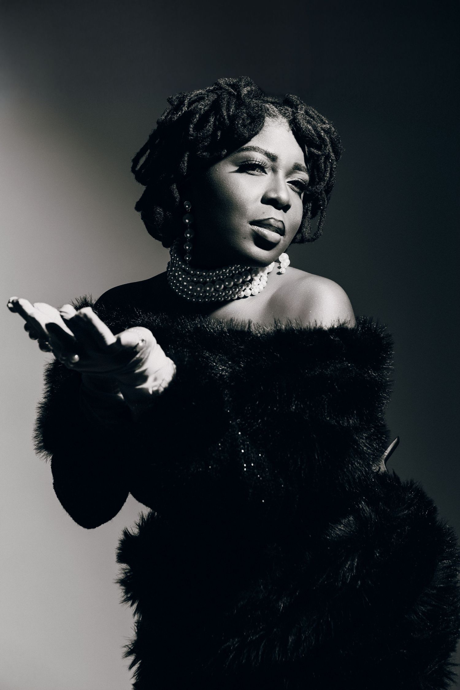 Black and white portrait of a woman in fur coat and pearl necklace, striking a confident pose with dramatic lighting.