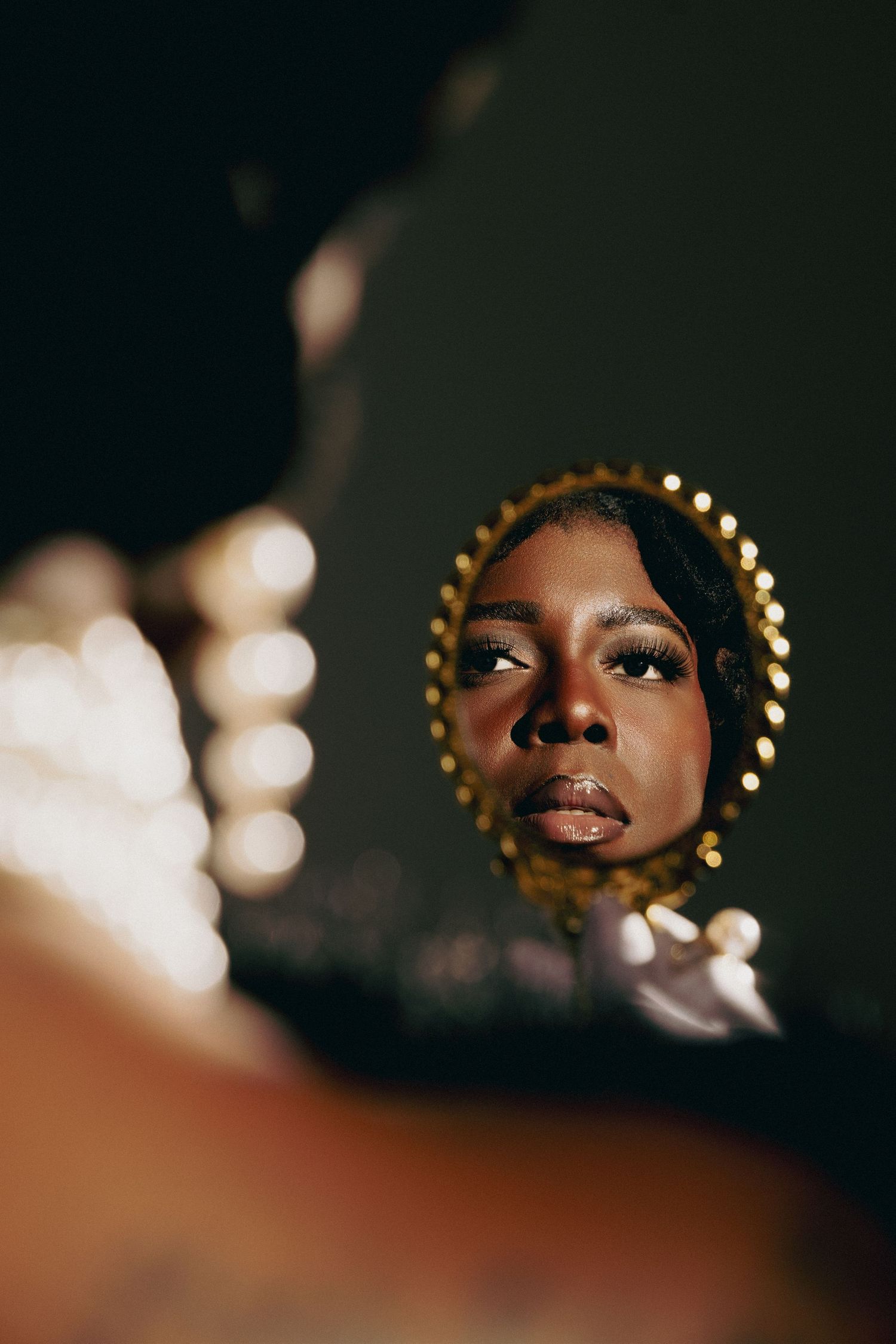 A Black woman with dramatic makeup and gold headpiece gazes into an ornate round mirror in moody lighting.