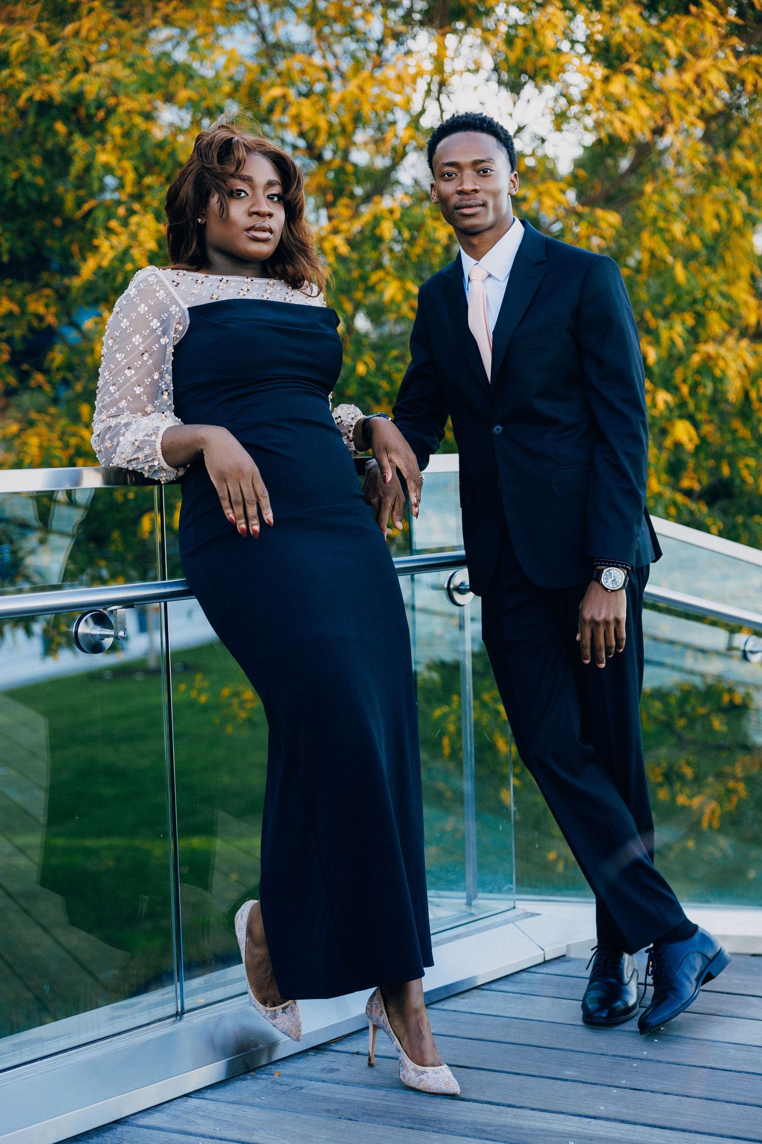 Elegant couple in formal black attire posing on a glass balcony surrounded by autumn foliage.