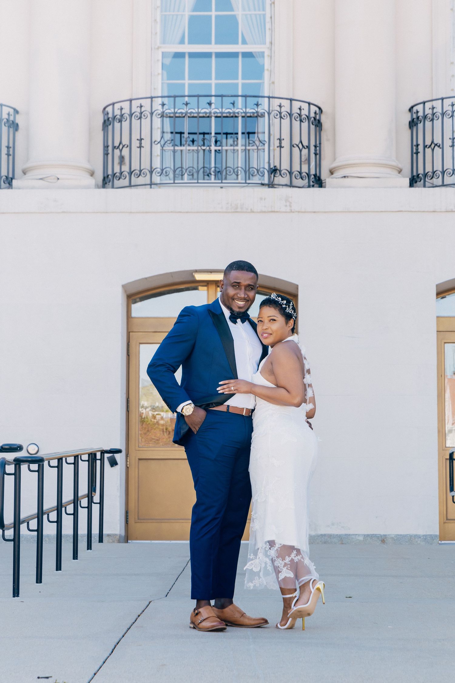 Newlywed couple poses together outside a classic white building, groom in navy suit and bride in lace gown.