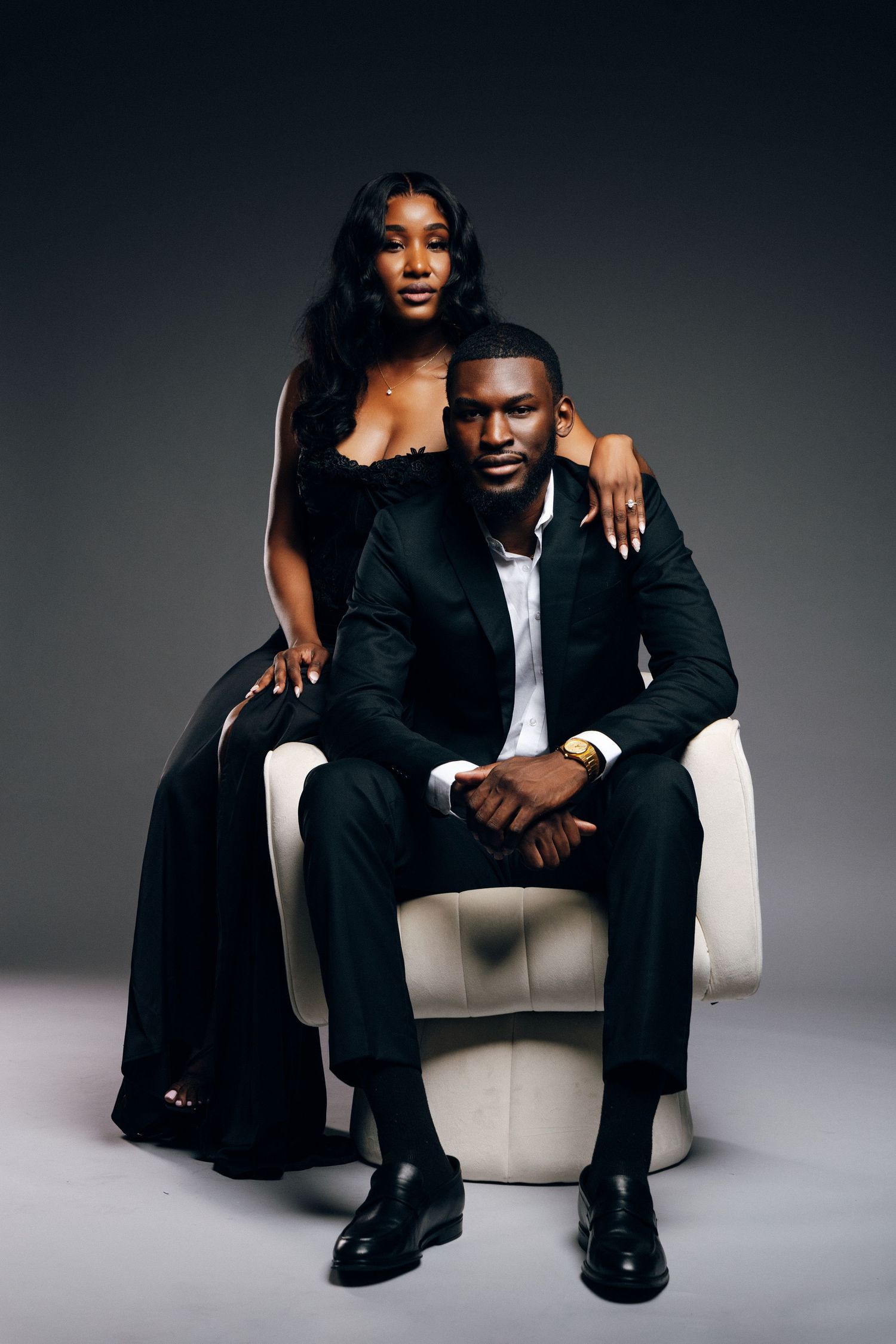 Elegant Black couple in matching black formal attire posing in studio, man seated in white chair, woman standing behind.