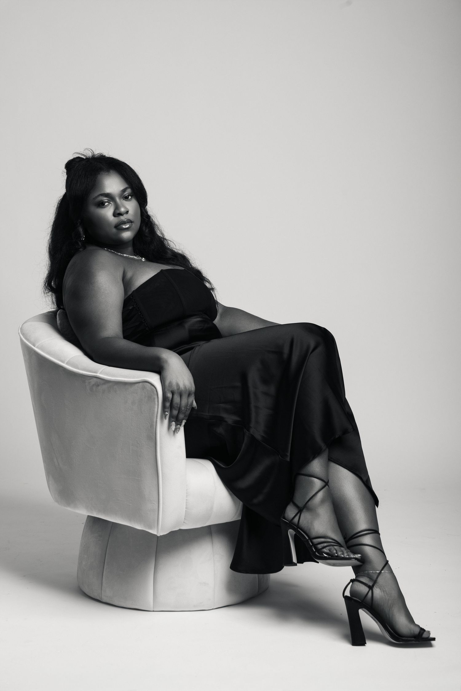 Black and white portrait of a woman in elegant black outfit seated gracefully in a white velvet chair.