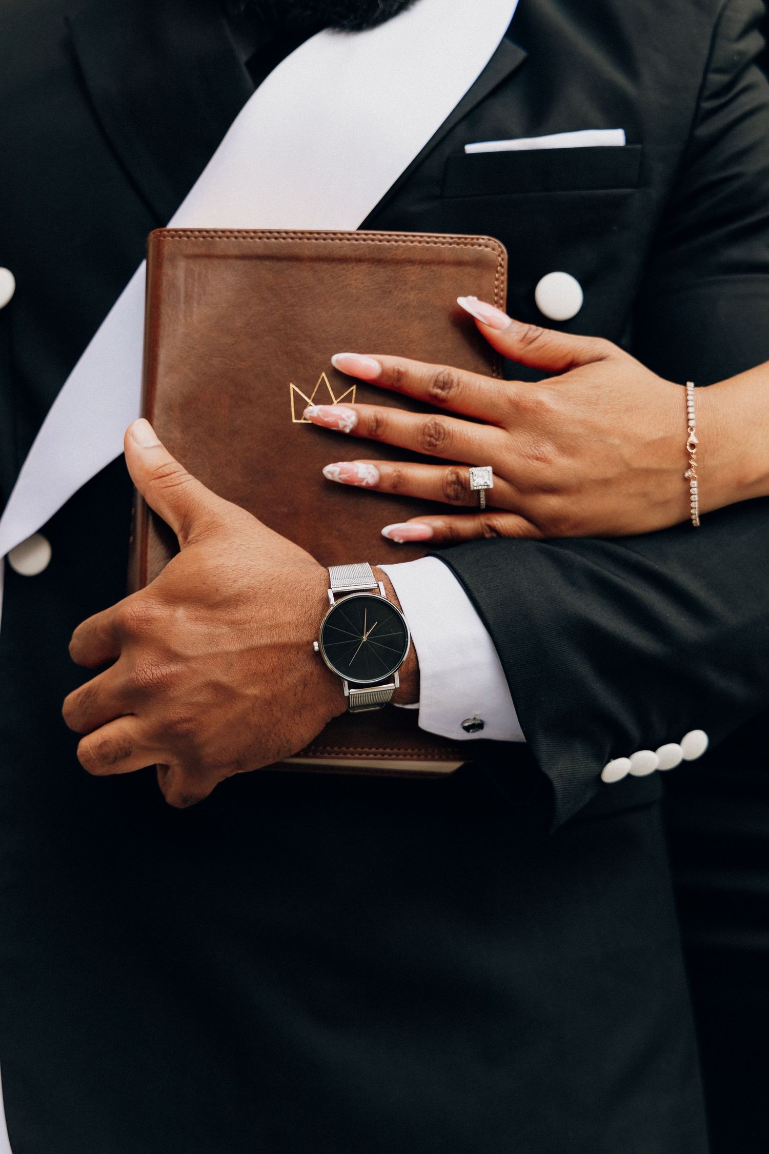 Couple holding a brown leather Bible, groom in black tuxedo with silver watch, bride with manicured nails and diamond ring.