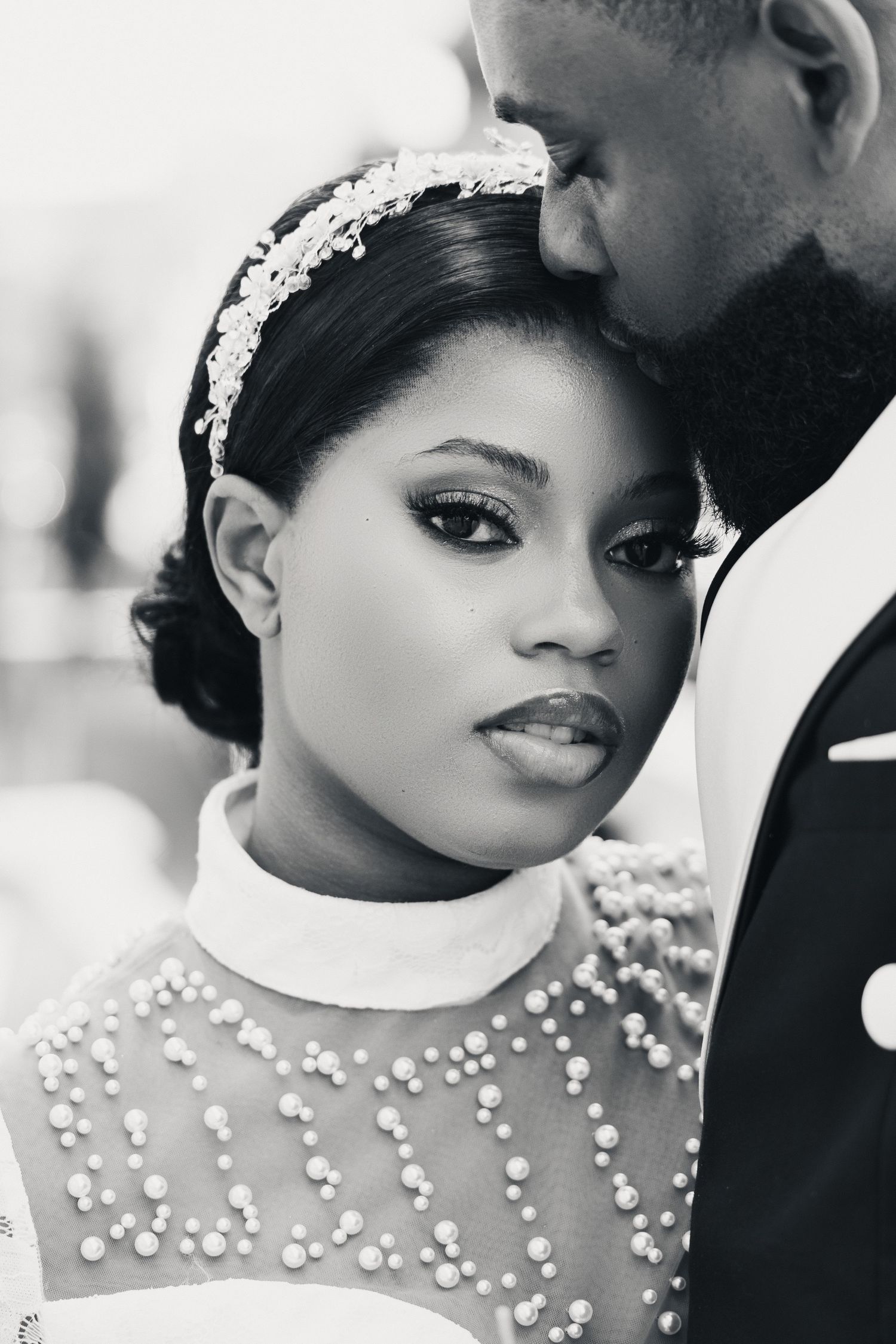 Black and white portrait of a bride in pearl-adorned dress with headpiece, groom kissing her forehead at wedding.
