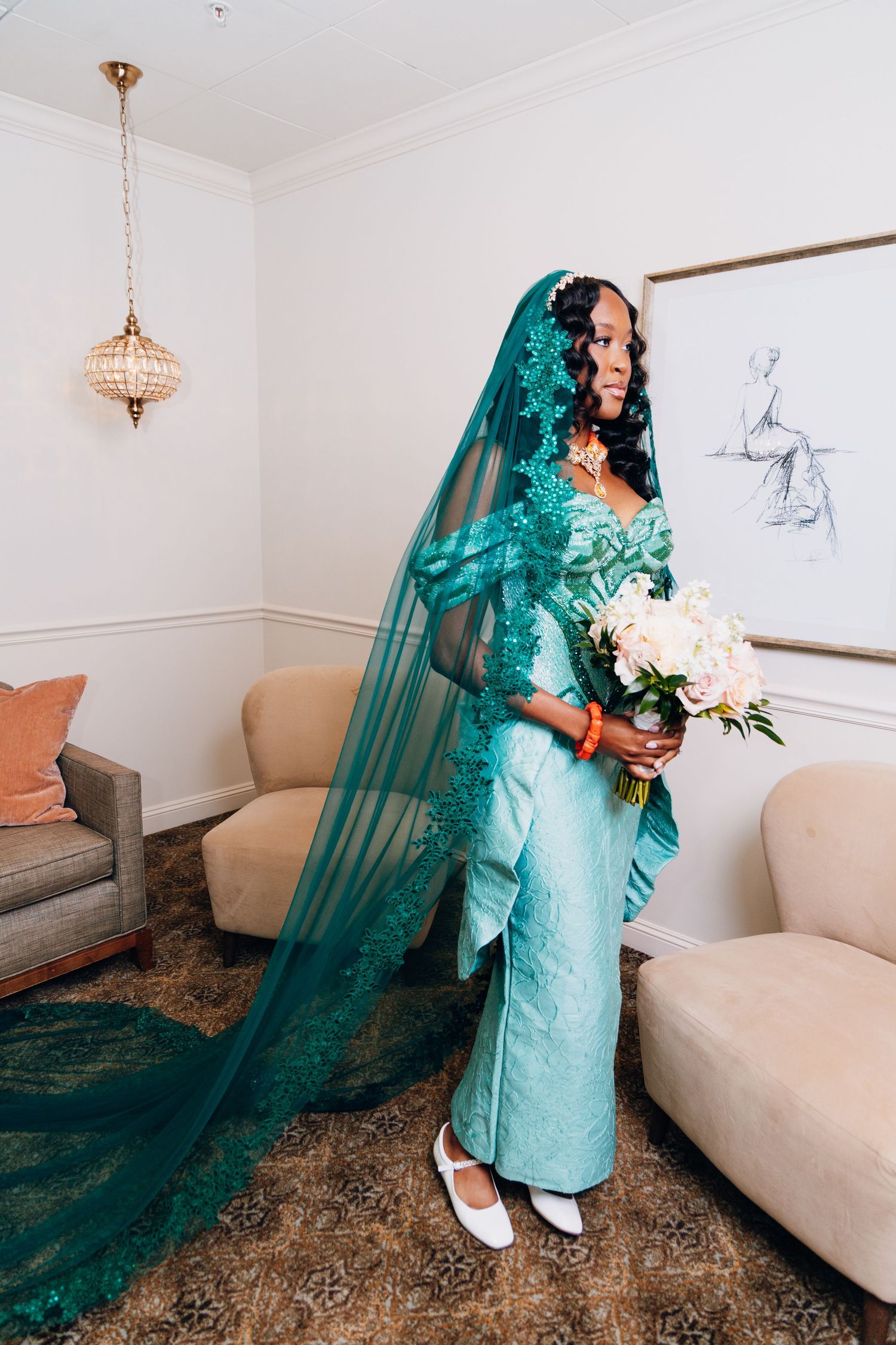 Bride in teal lace gown and matching veil holds white bouquet in elegant room.