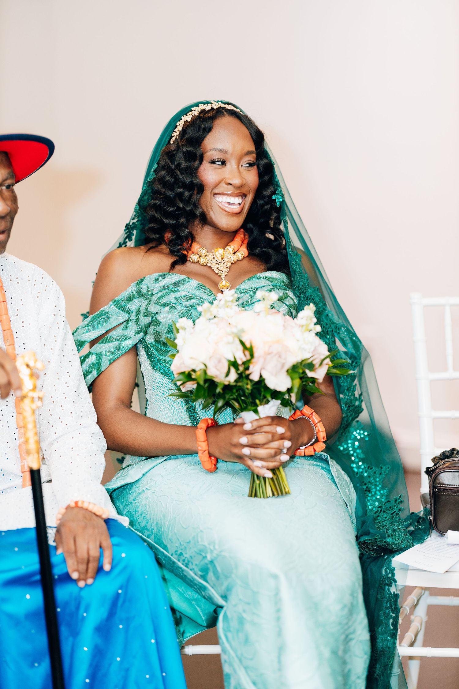 Radiant bride in teal African lace gown and veil smiles while holding white rose bouquet at traditional ceremony.