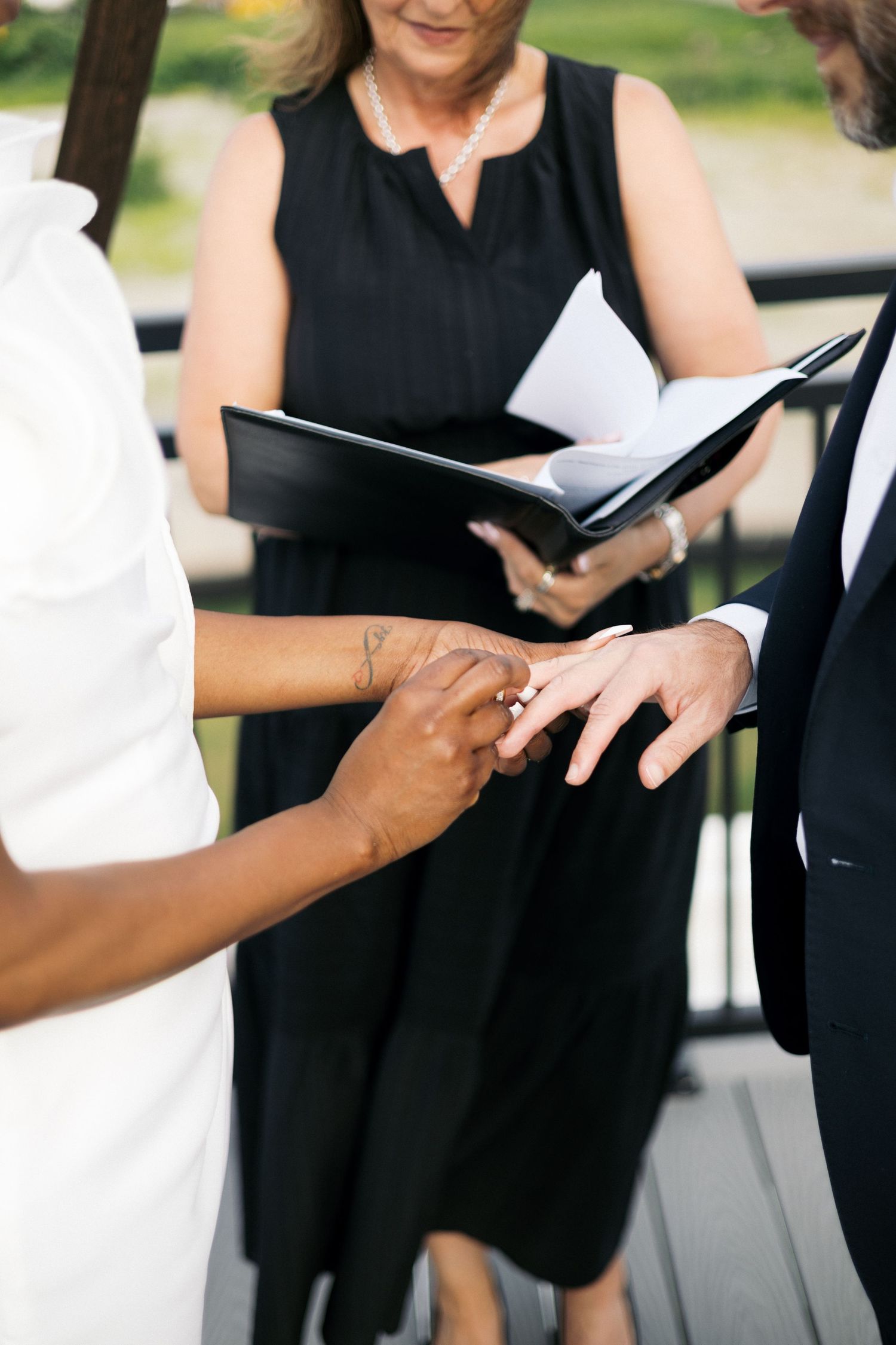 Couple exchanging rings during outdoor wedding ceremony with officiant holding book in background.