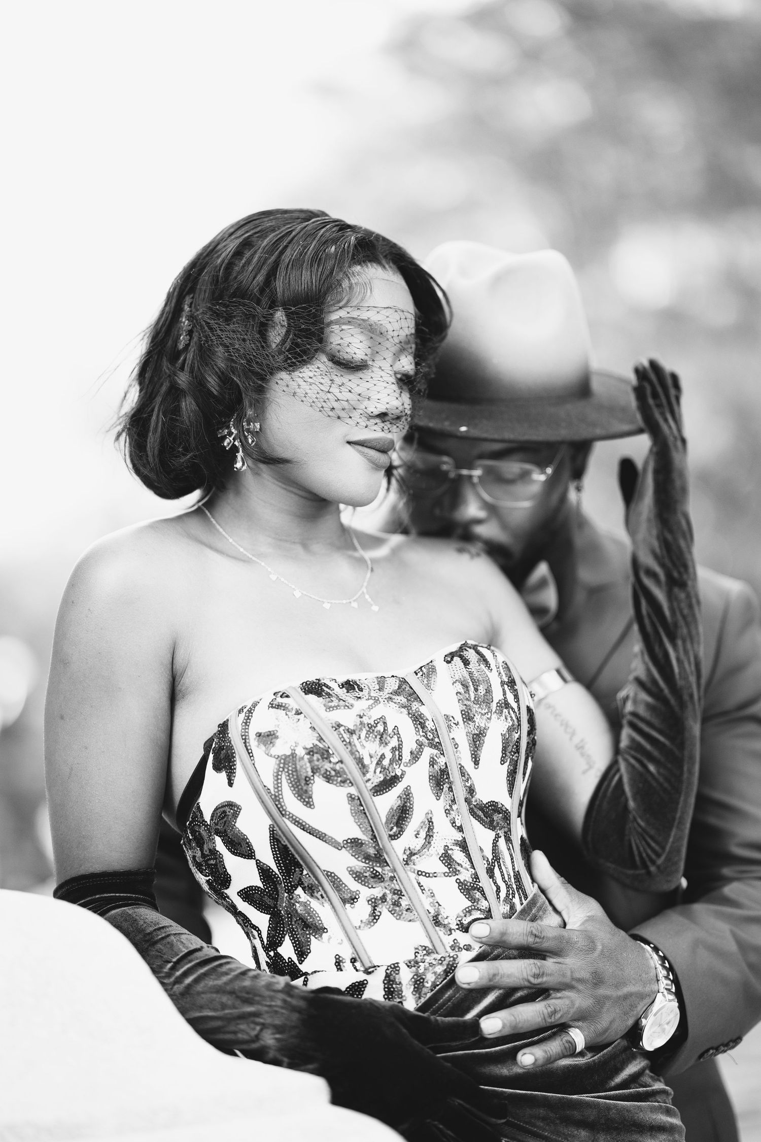 Black and white portrait of a glamorous couple, woman in corset with veil, man in hat embracing her.
