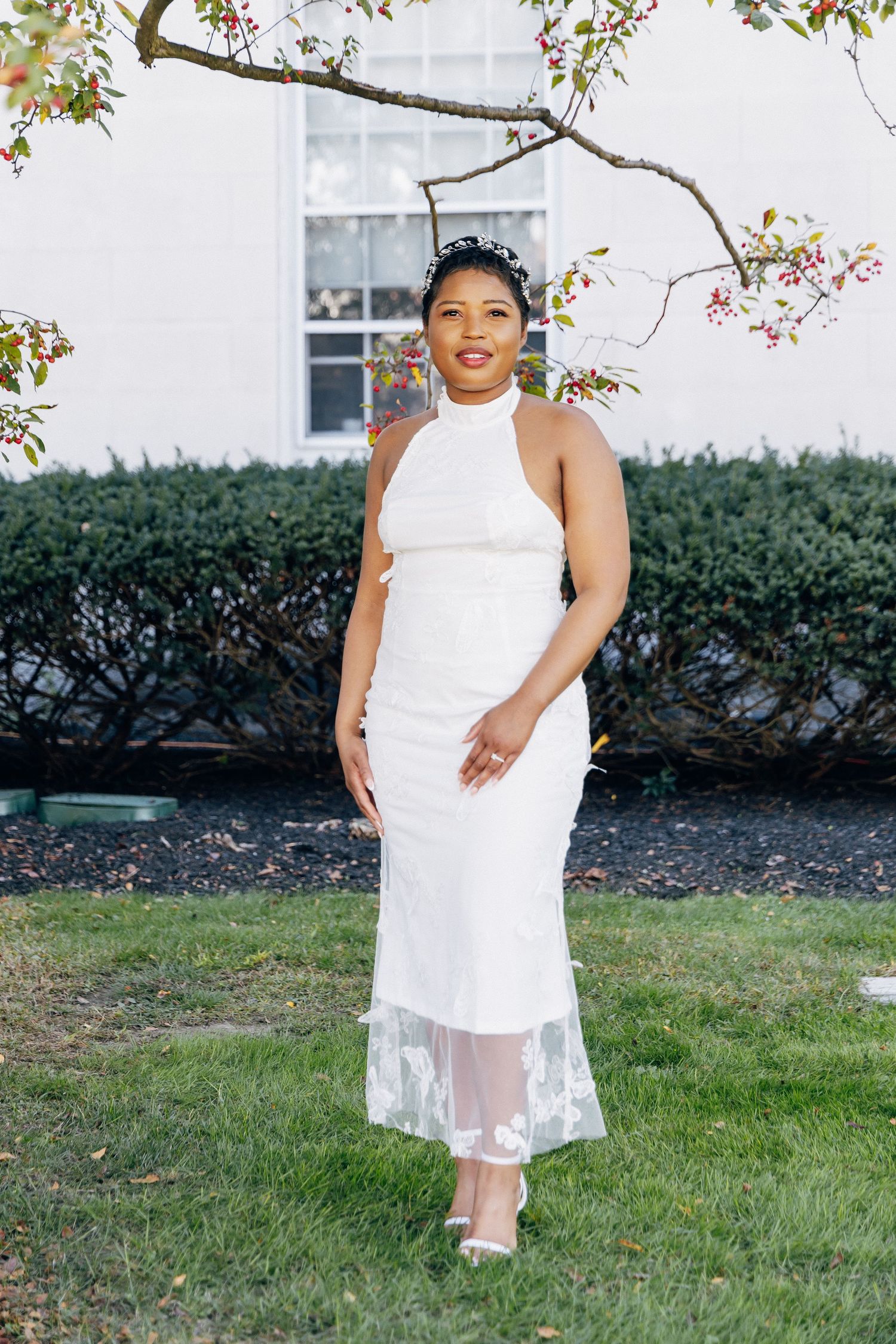 Bride poses outdoors in white halter lace midi dress with floral headband under blooming spring tree branches.