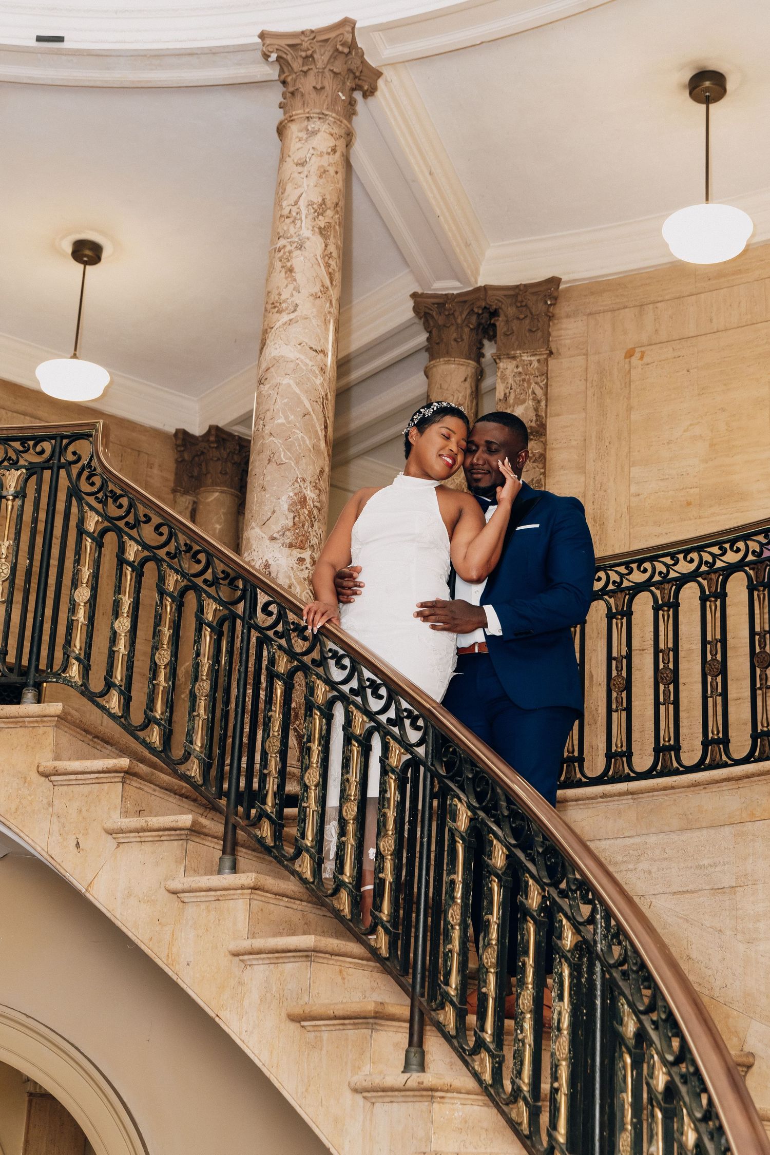 Bride and groom pose romantically on elegant curved staircase with ornate iron railing in grand marble venue.