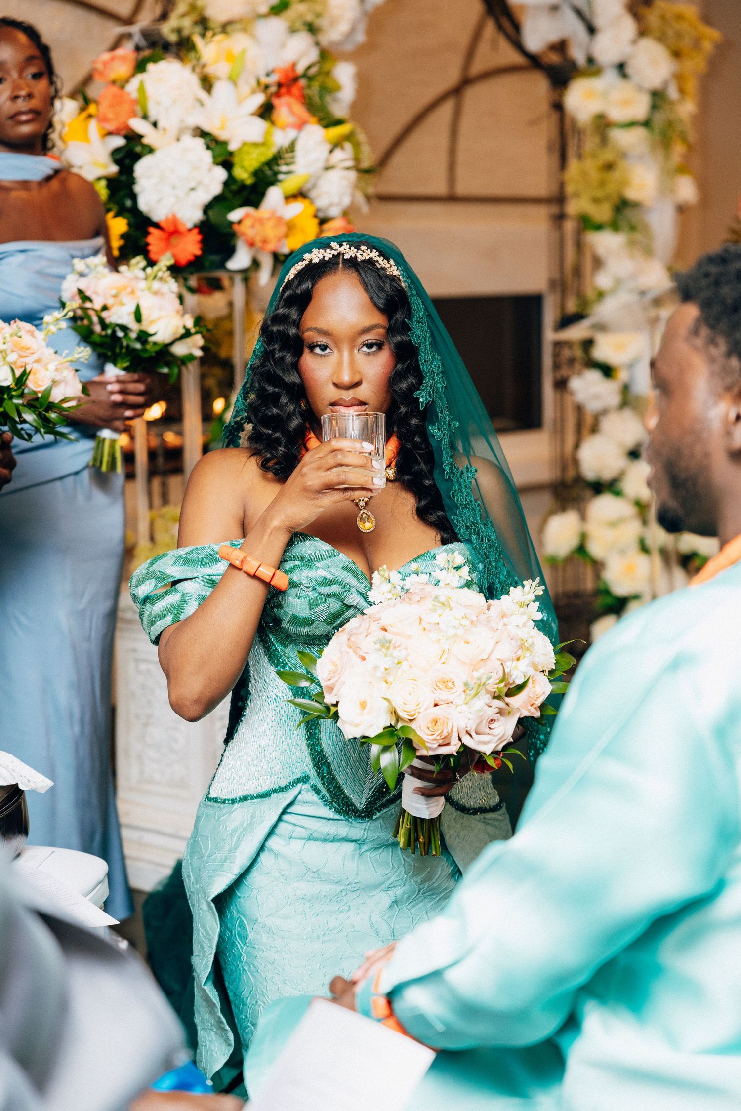 Bride in teal African lace gown sips from glass during traditional Nigerian wedding ceremony with floral backdrop.