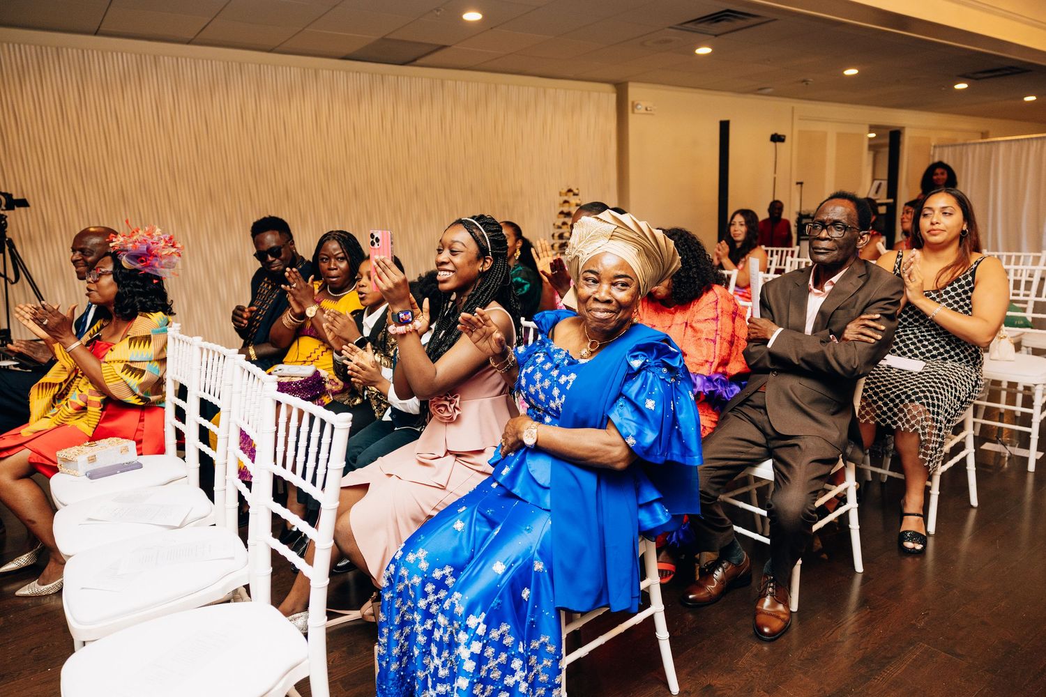 Guests in colorful traditional African attire clapping and celebrating at an indoor event or ceremony.