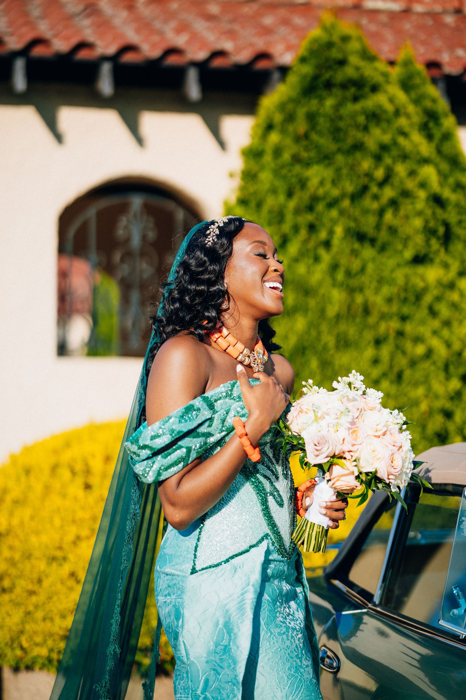 Joyful bride in teal African wedding dress with coral jewelry holding bouquet beside classic car.
