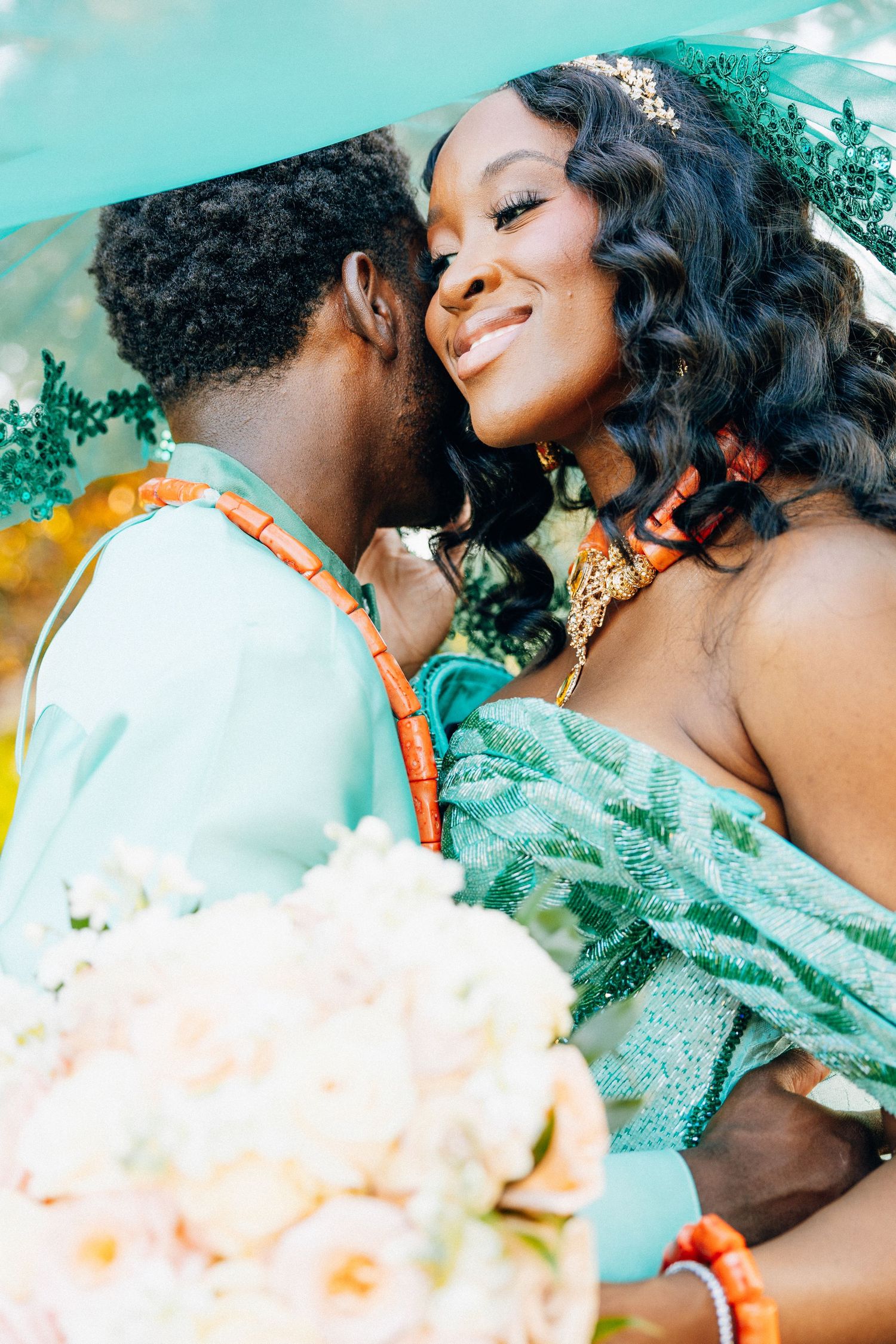 Nigerian bride and groom in matching teal outfits share romantic moment under green veil outdoors.