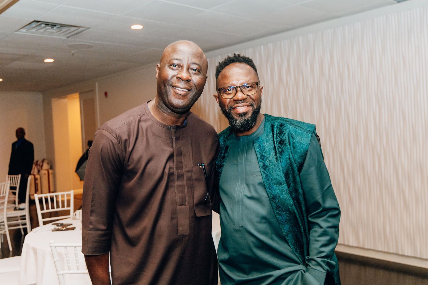 Two men in traditional African attire smiling together at a wedding reception venue.