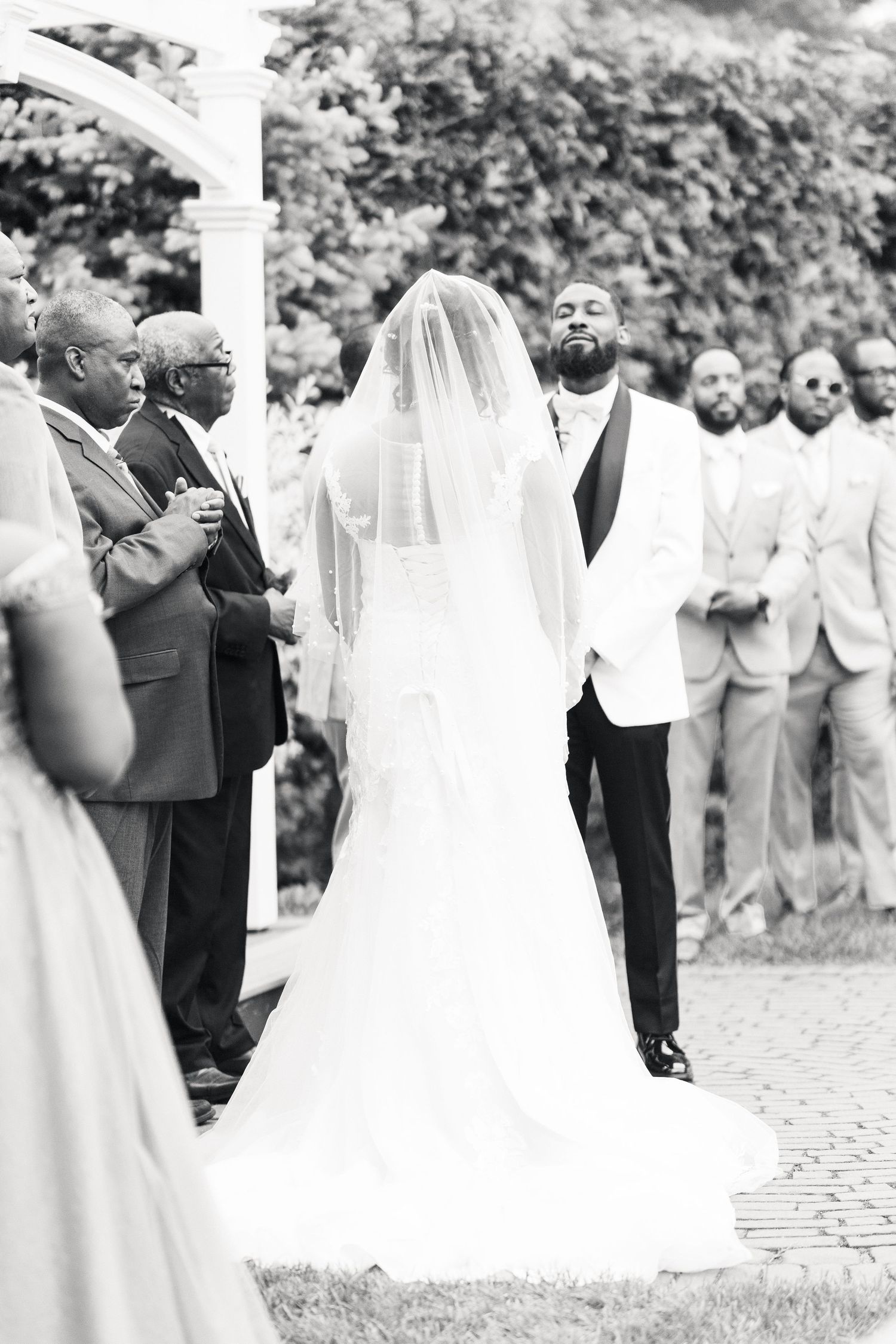 Groom looks up emotionally as veiled bride approaches outdoor wedding altar in black and white photo.