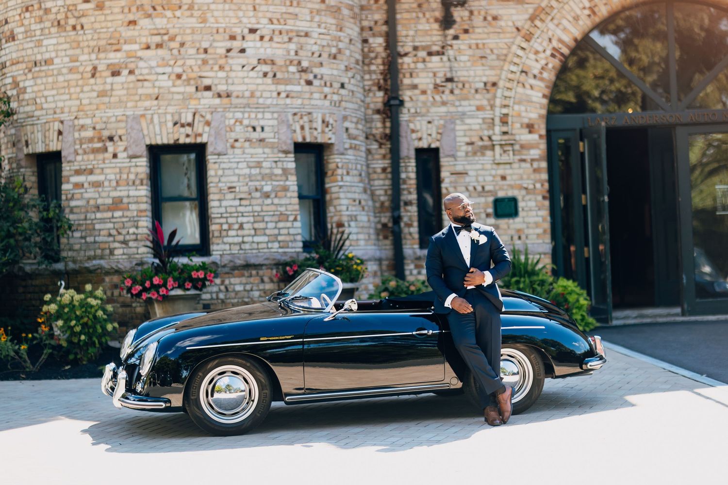 Dapper groom in navy tuxedo with bow tie leans against a classic black Porsche 356 outside brick building.