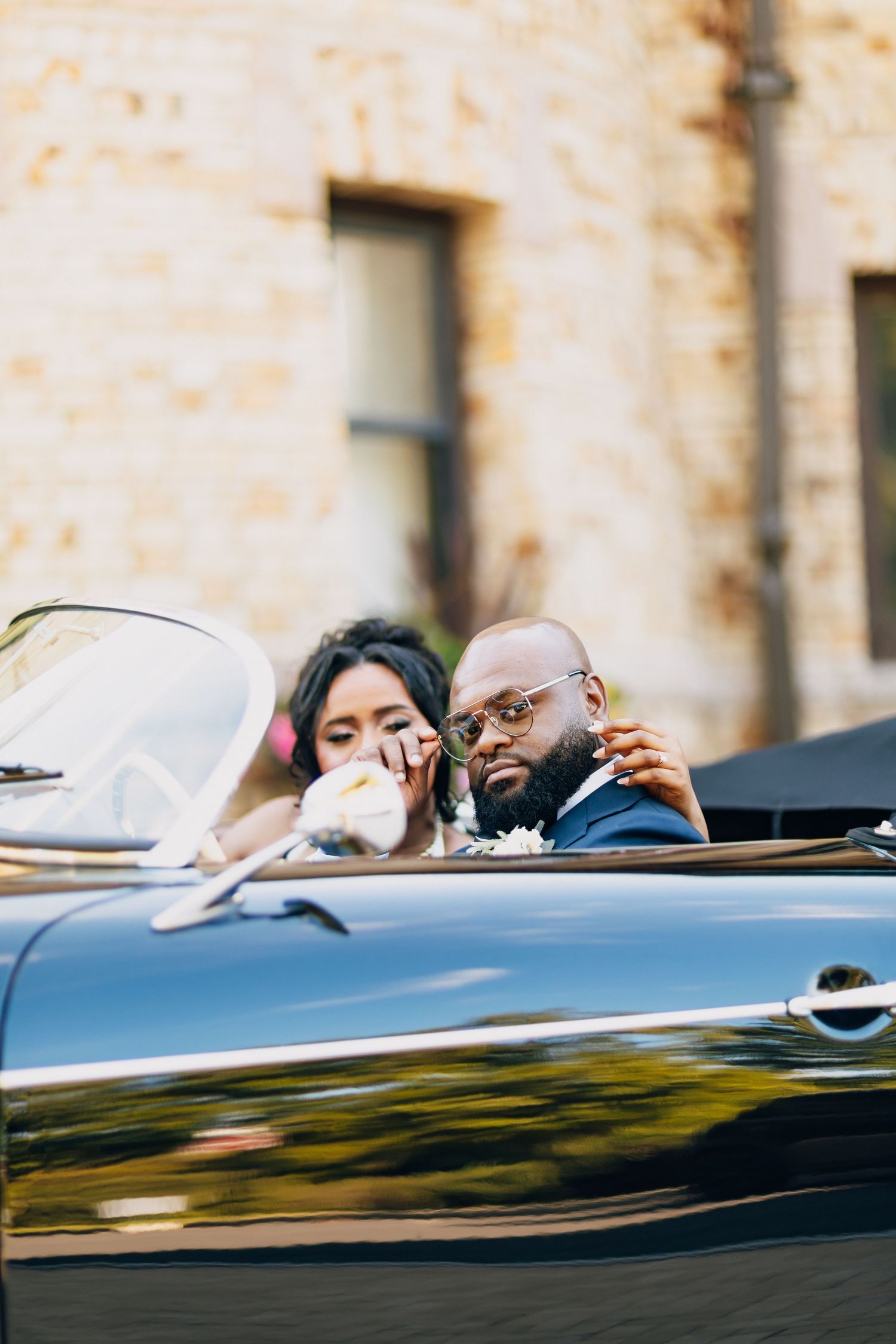 Bride and groom sharing an intimate moment in a vintage black convertible beside a stone building.