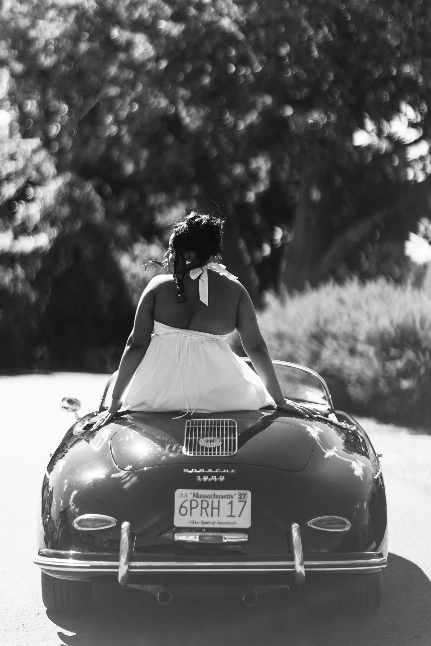 Black and white rear view of bride in halter dress leaning on vintage Porsche convertible trunk.