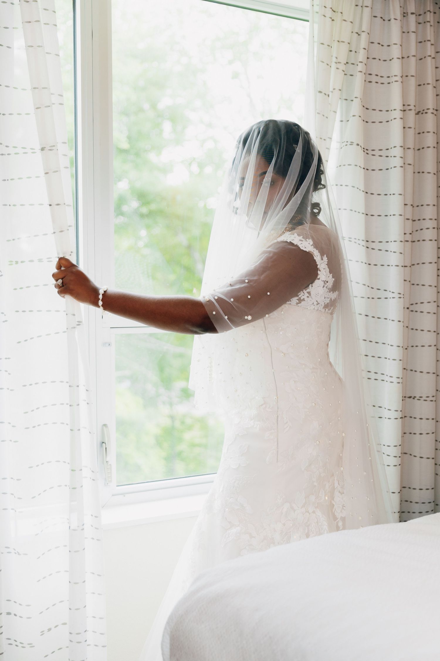 Bride in lace wedding dress and veil stands by bright window, looking outside on her wedding day.