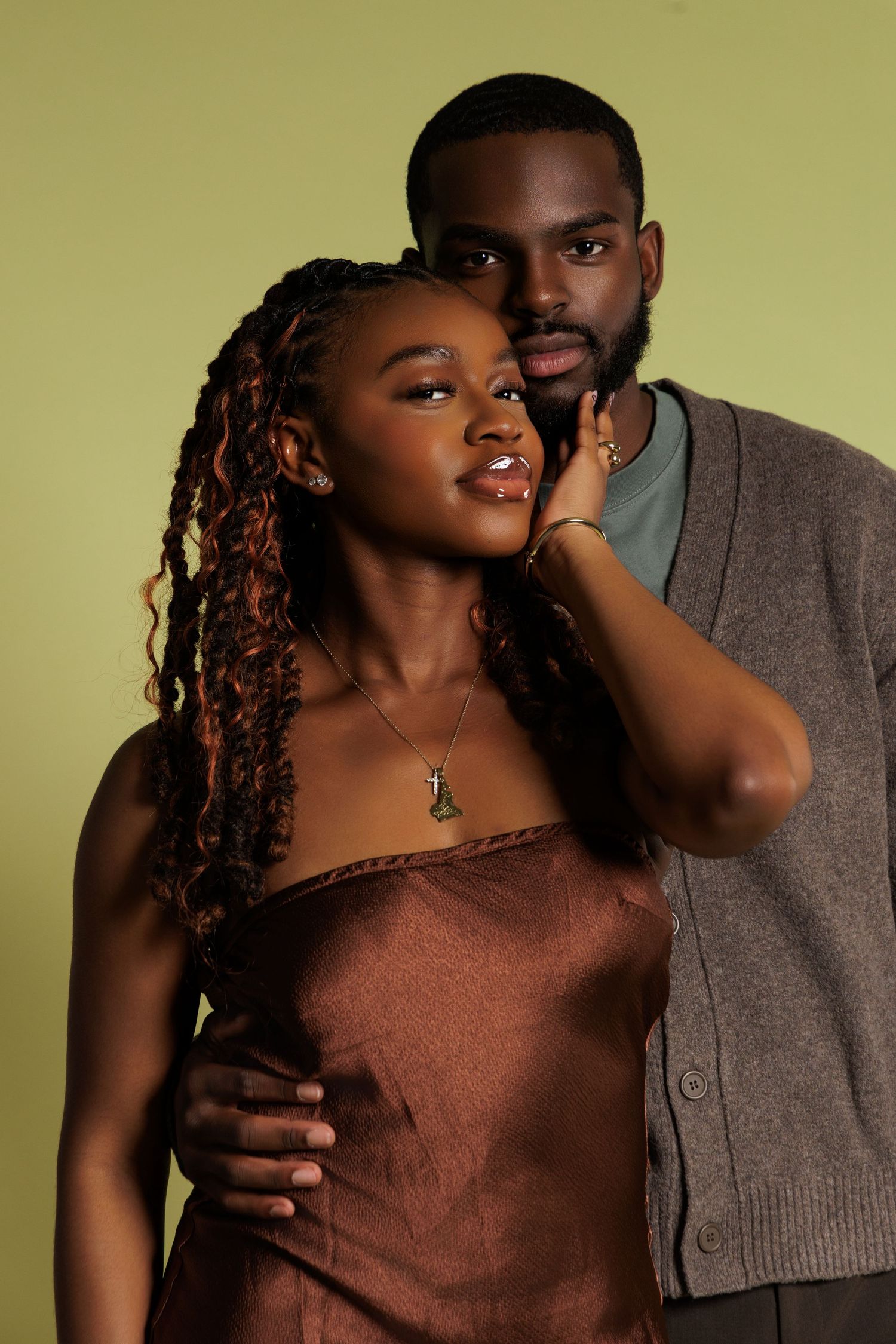 A Black couple poses together against a green background, woman in brown satin dress with braids, man in gray cardigan.