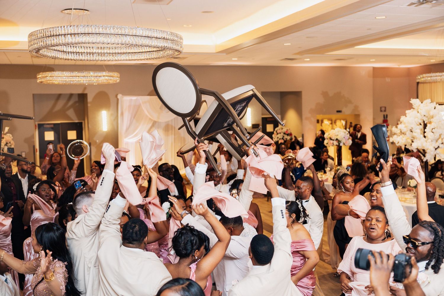 Wedding guests celebrate energetically raising chairs and hands in the air at a lively reception party.