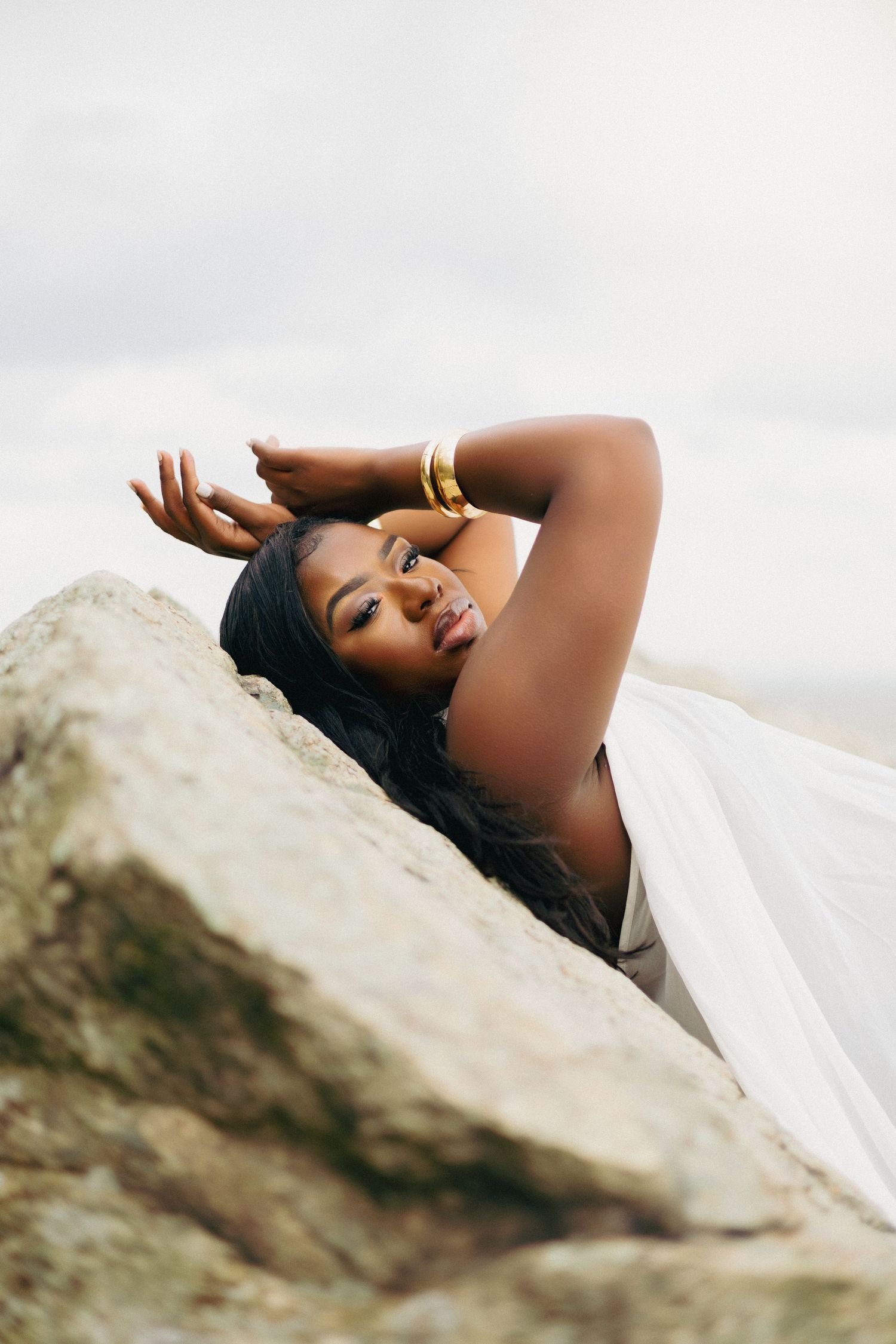 A Black woman in a white dress and gold bangles poses gracefully on large rocks against an overcast sky.