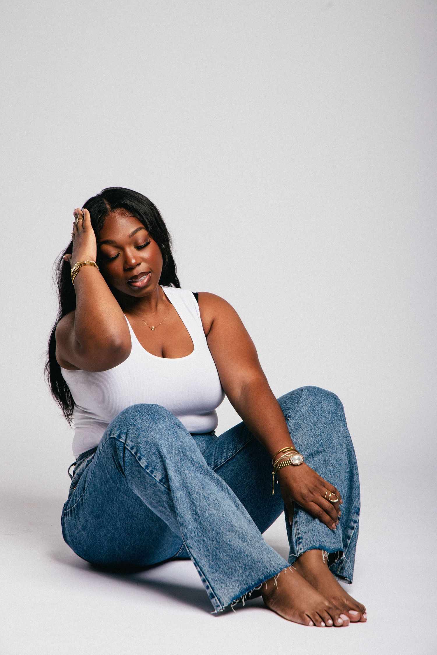 Woman in white tank top and blue jeans sitting cross-legged on white background with gold jewelry.