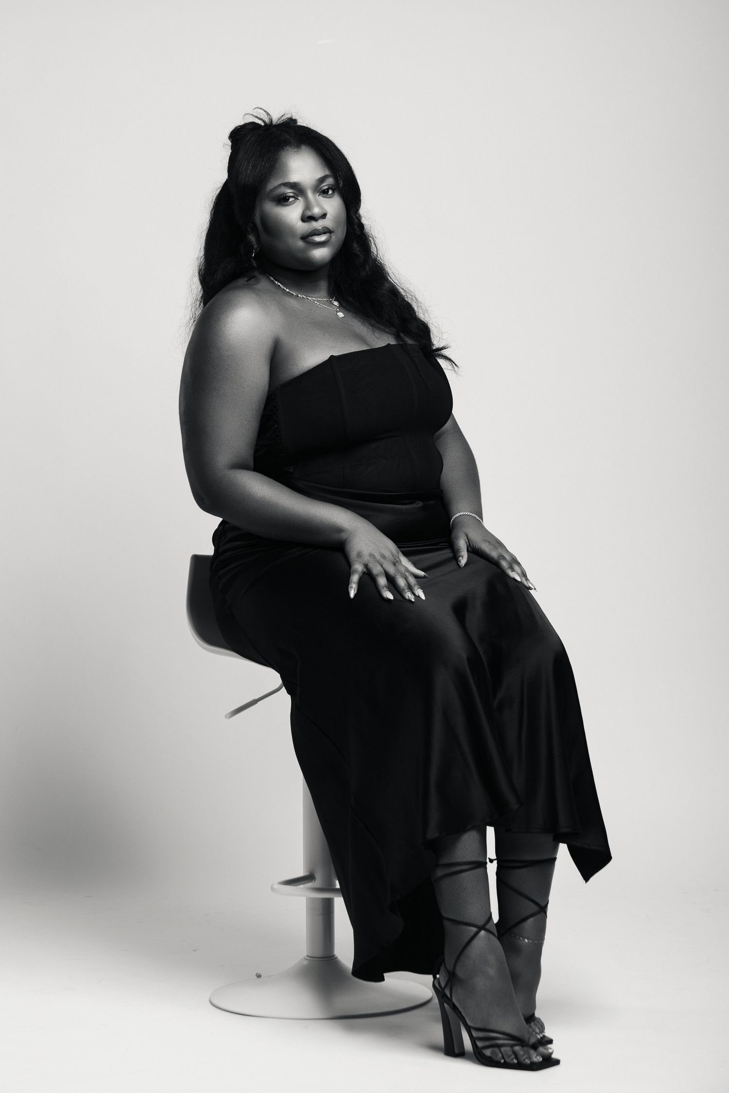 Black and white portrait of a woman in a black strapless dress seated on a stool, posing confidently.