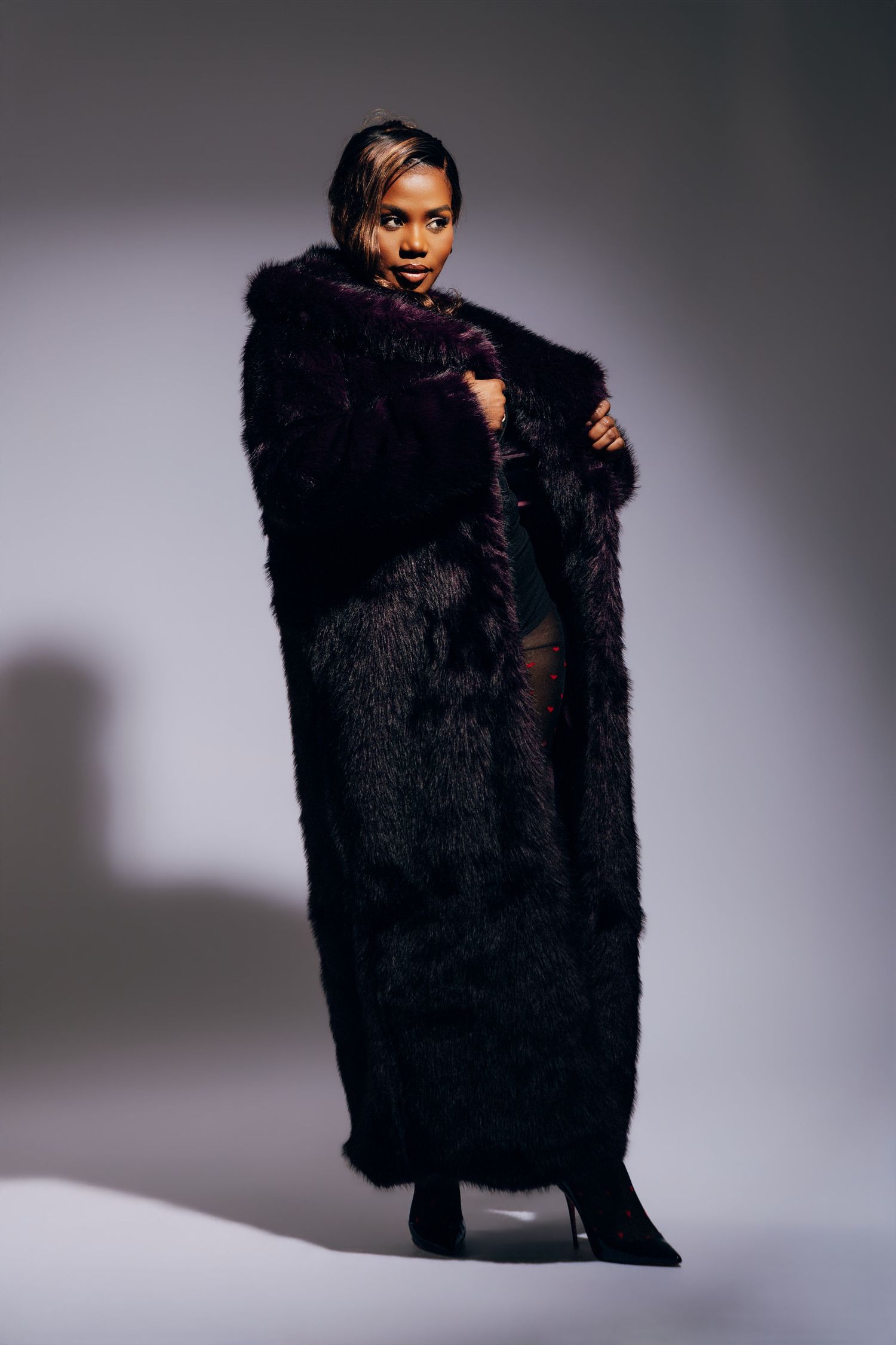 Woman in long black fur coat posing confidently in studio setting wearing black heels.