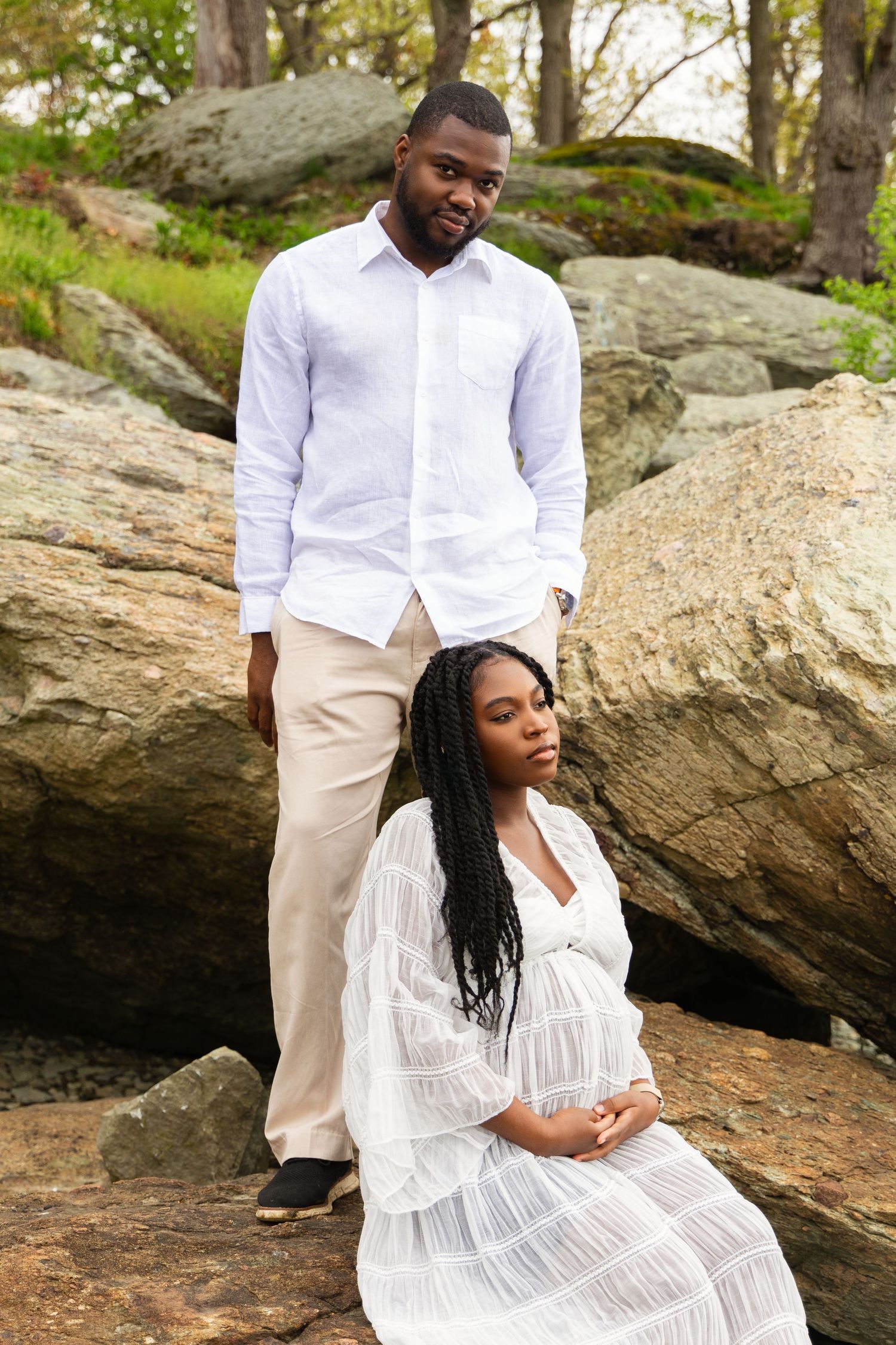 A pregnant woman in a white dress sits on rocks while a man in a white shirt stands behind her outdoors.