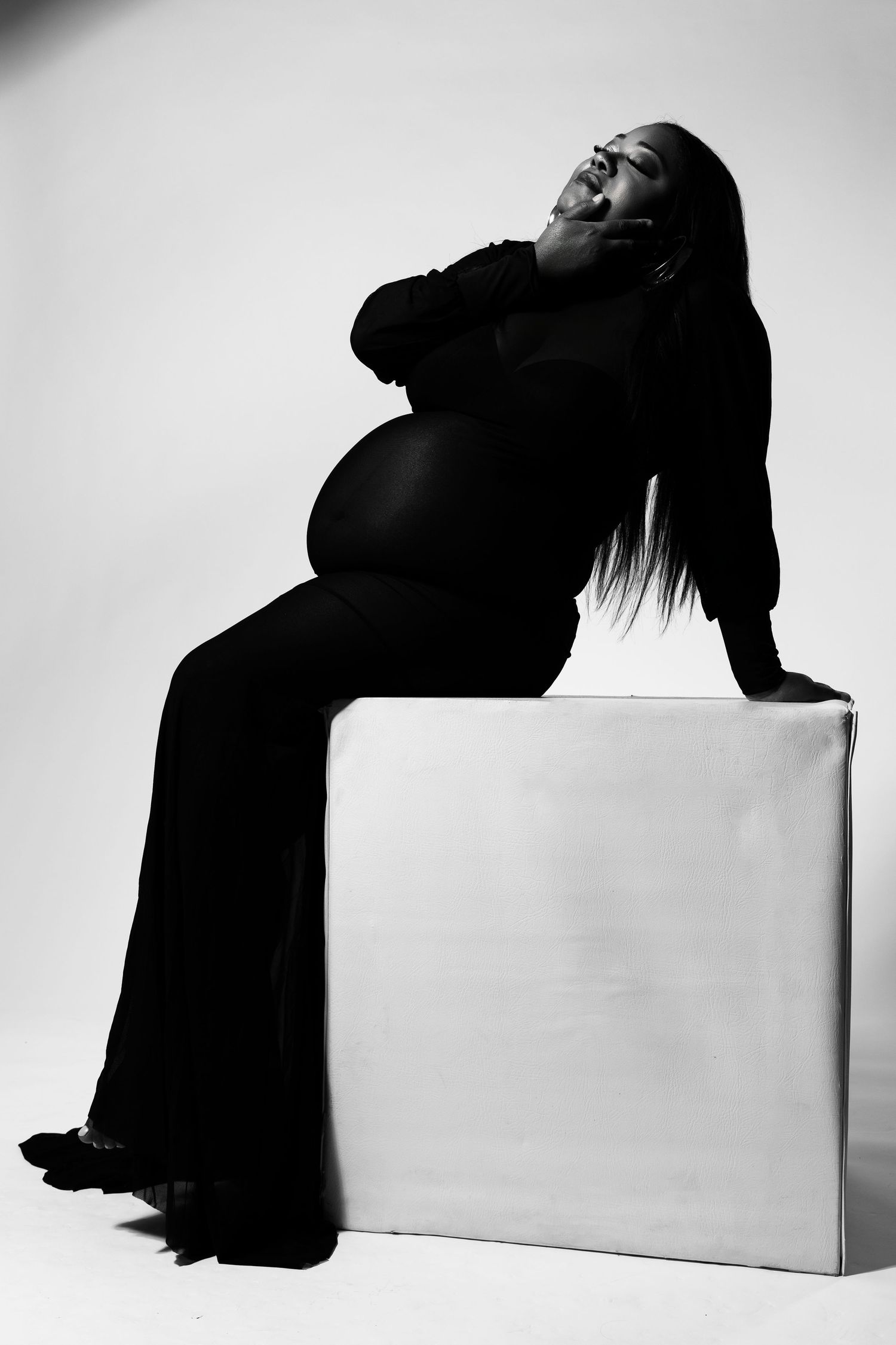 Black and white portrait of a pregnant woman in a black dress sitting on a white cube, posing gracefully.