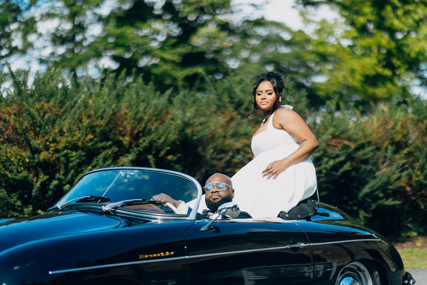 Bride in white dress stands behind groom seated in classic black convertible surrounded by lush green trees.