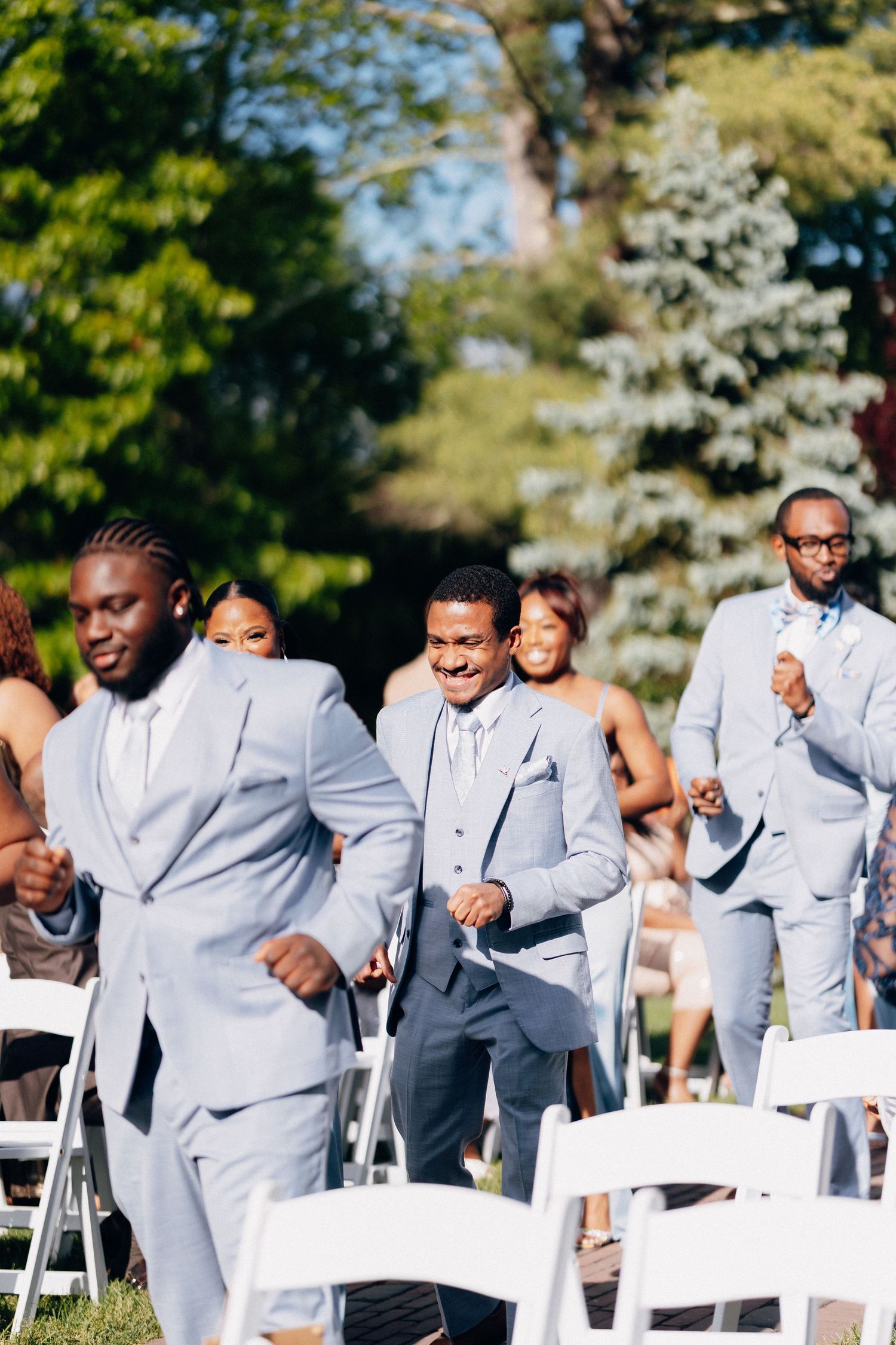 Groomsmen in light gray suits dancing joyfully down the outdoor wedding aisle lined with white chairs and lush greenery.