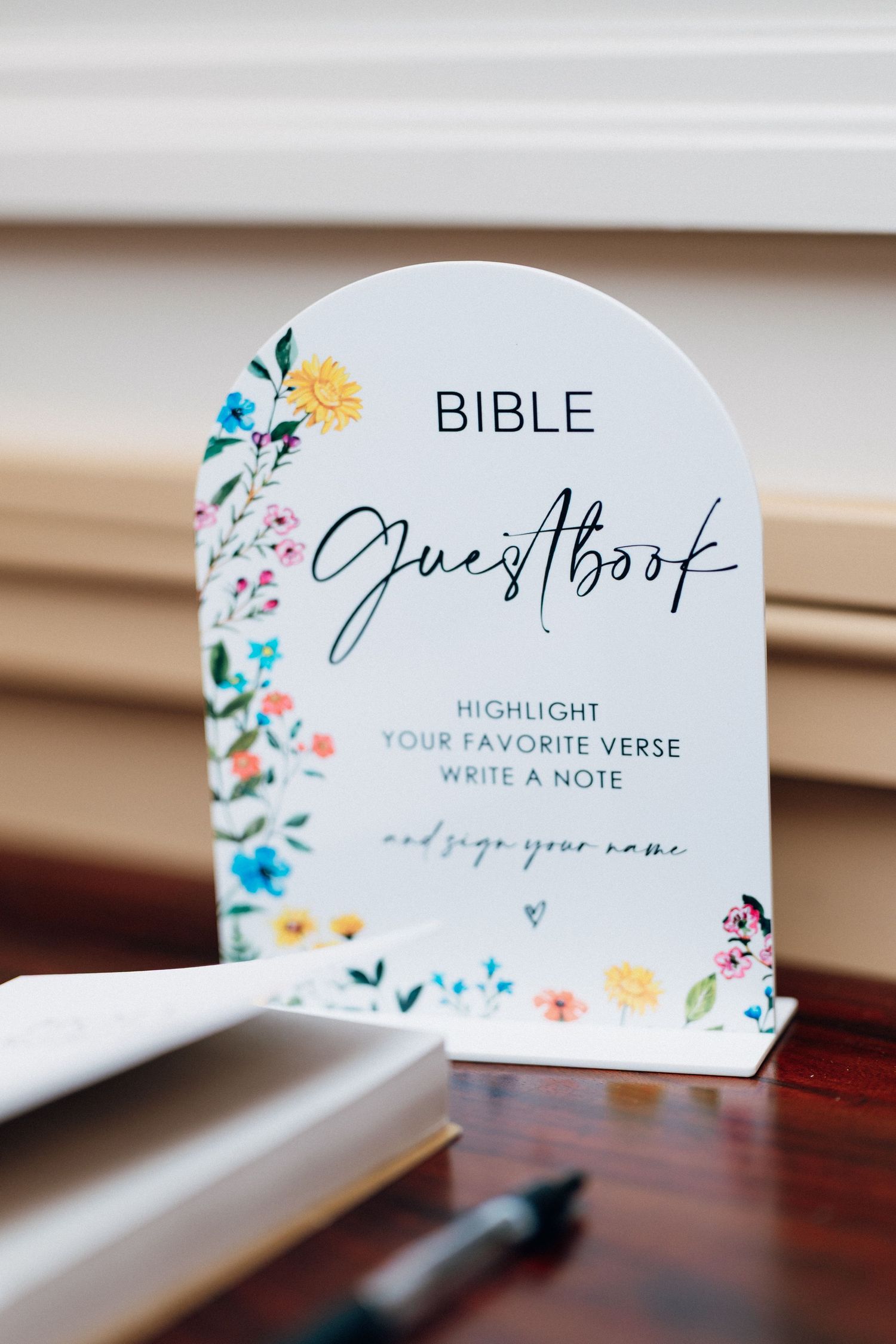 Arch-shaped Bible guestbook sign with colorful wildflowers inviting guests to highlight a verse and sign.