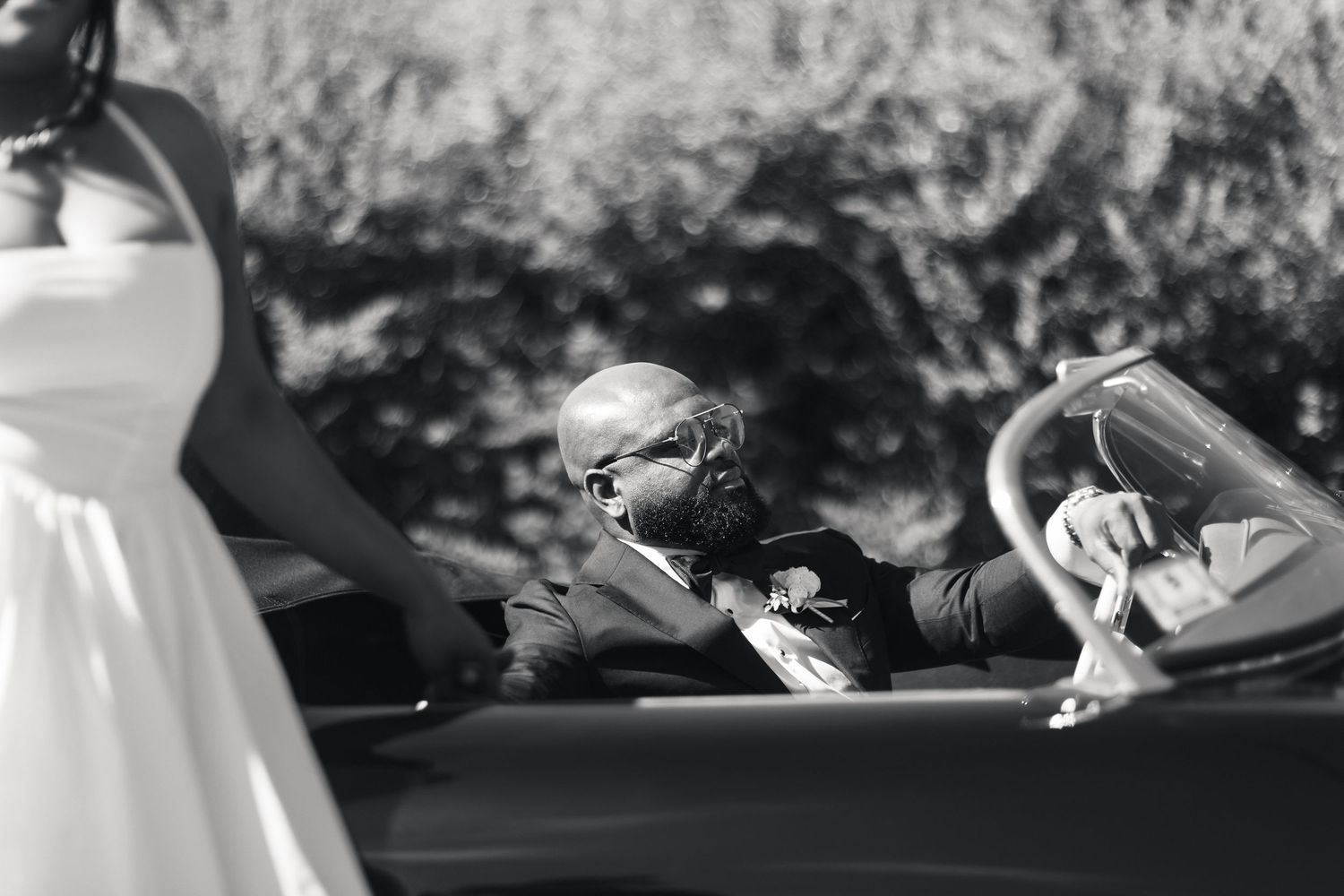 Black and white photo of groom leaning from vintage convertible toward bride on wedding day.