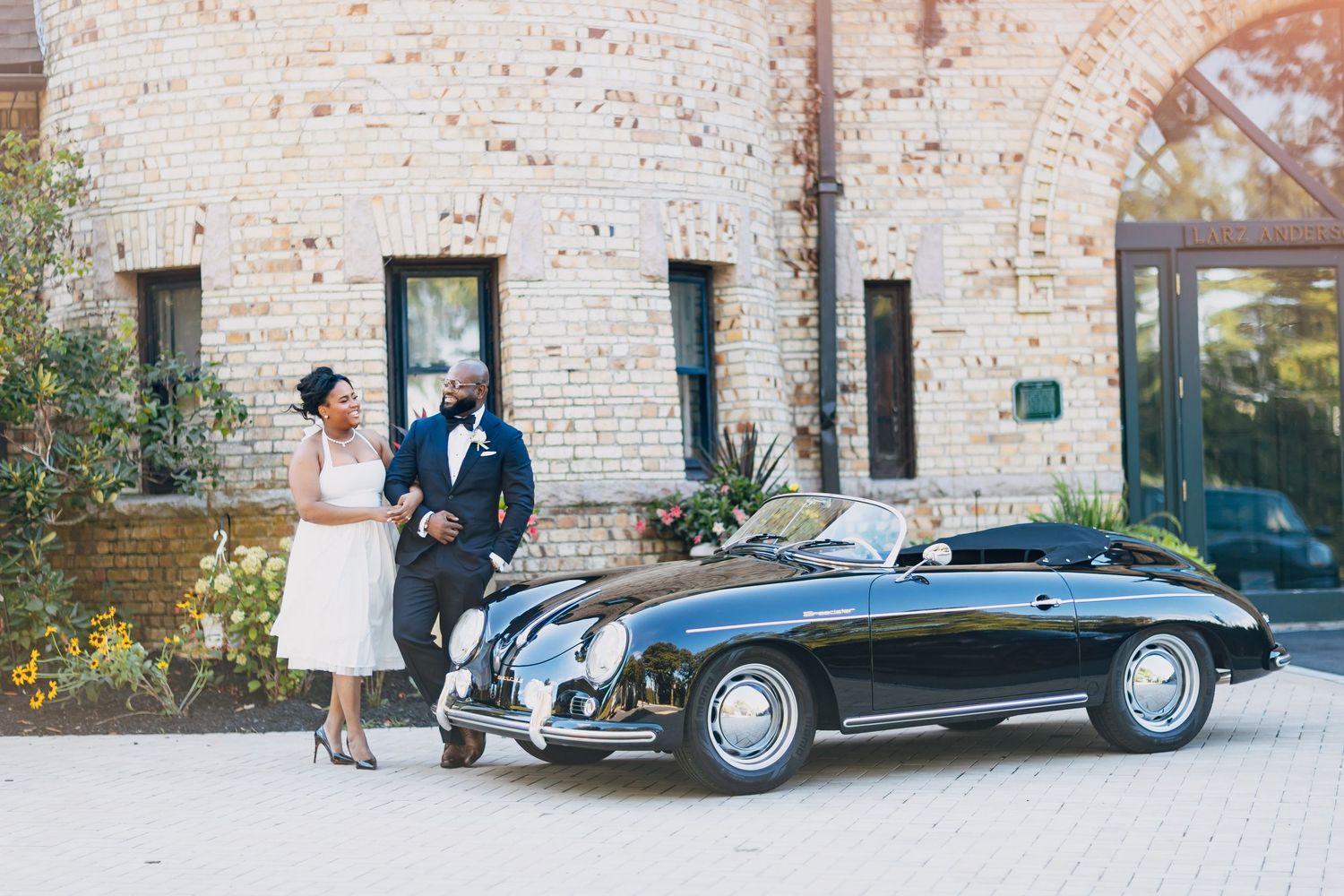 Bride and groom in wedding attire posing beside a classic black vintage Porsche 356 convertible outside a brick building.