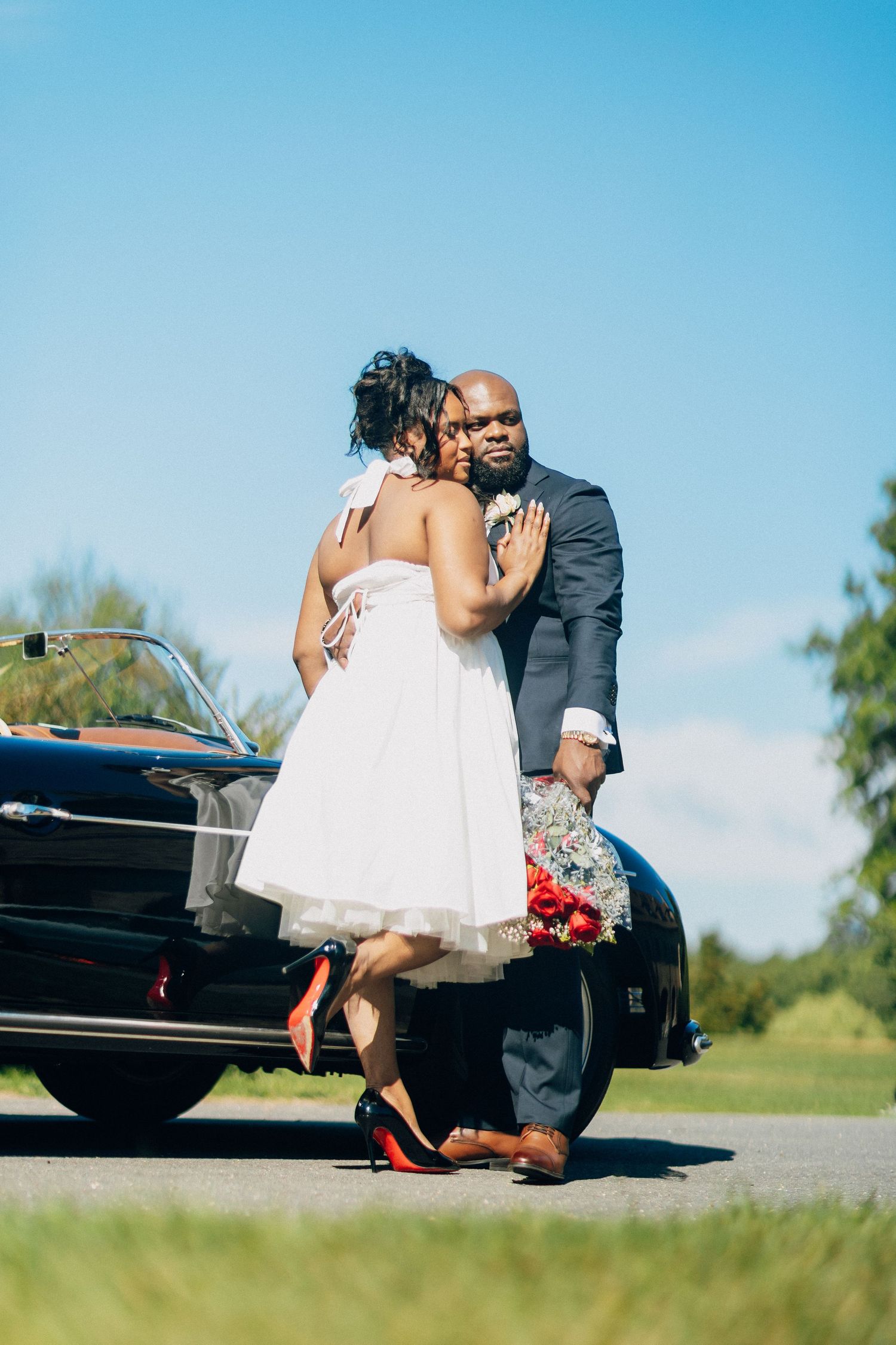 Bride in white dress and red heels embraces groom in navy suit beside a classic black convertible.