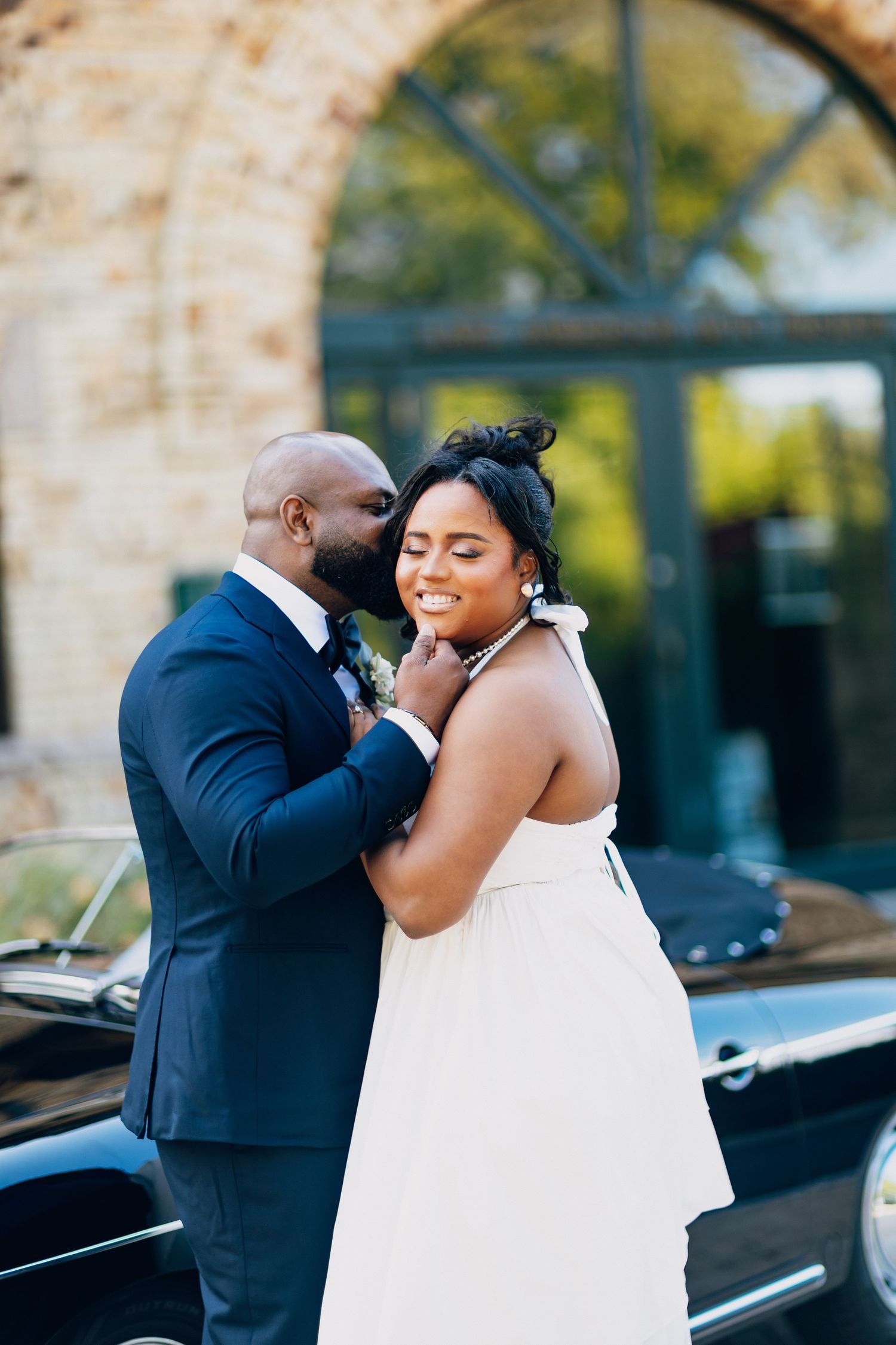 Groom in navy suit tenderly holds bride's face as she smiles in a white halter wedding gown.