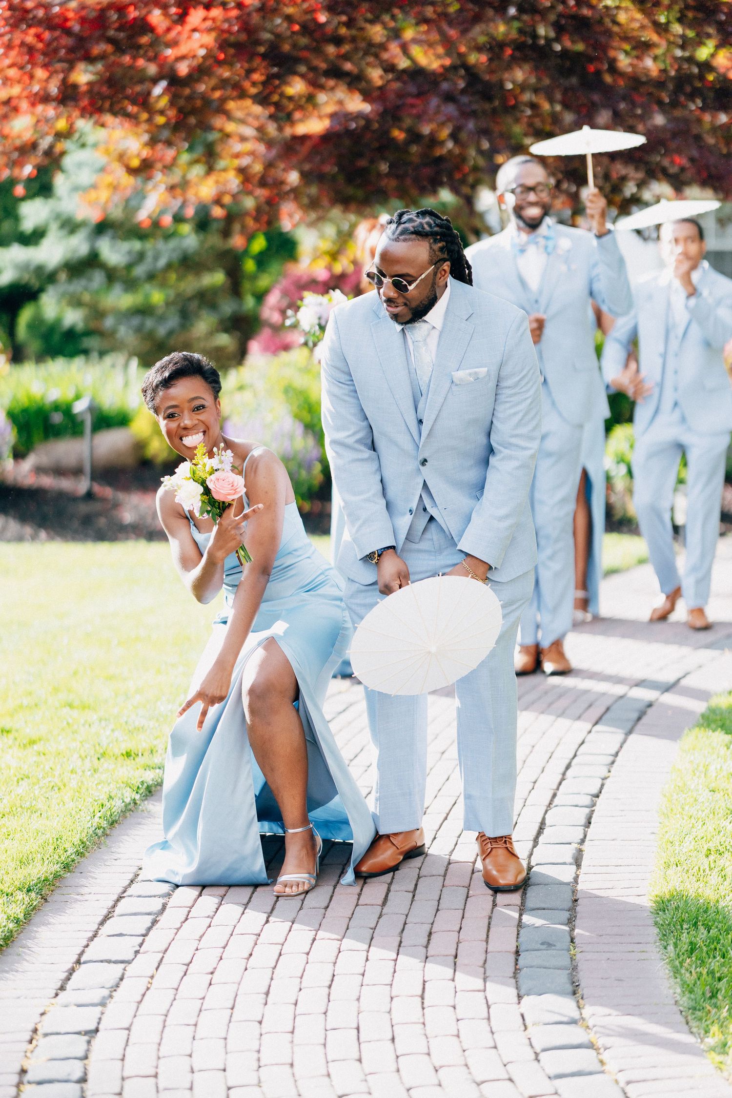 A bridesmaid in a light blue gown and groomsman in a matching suit walk together during a sunny outdoor wedding.