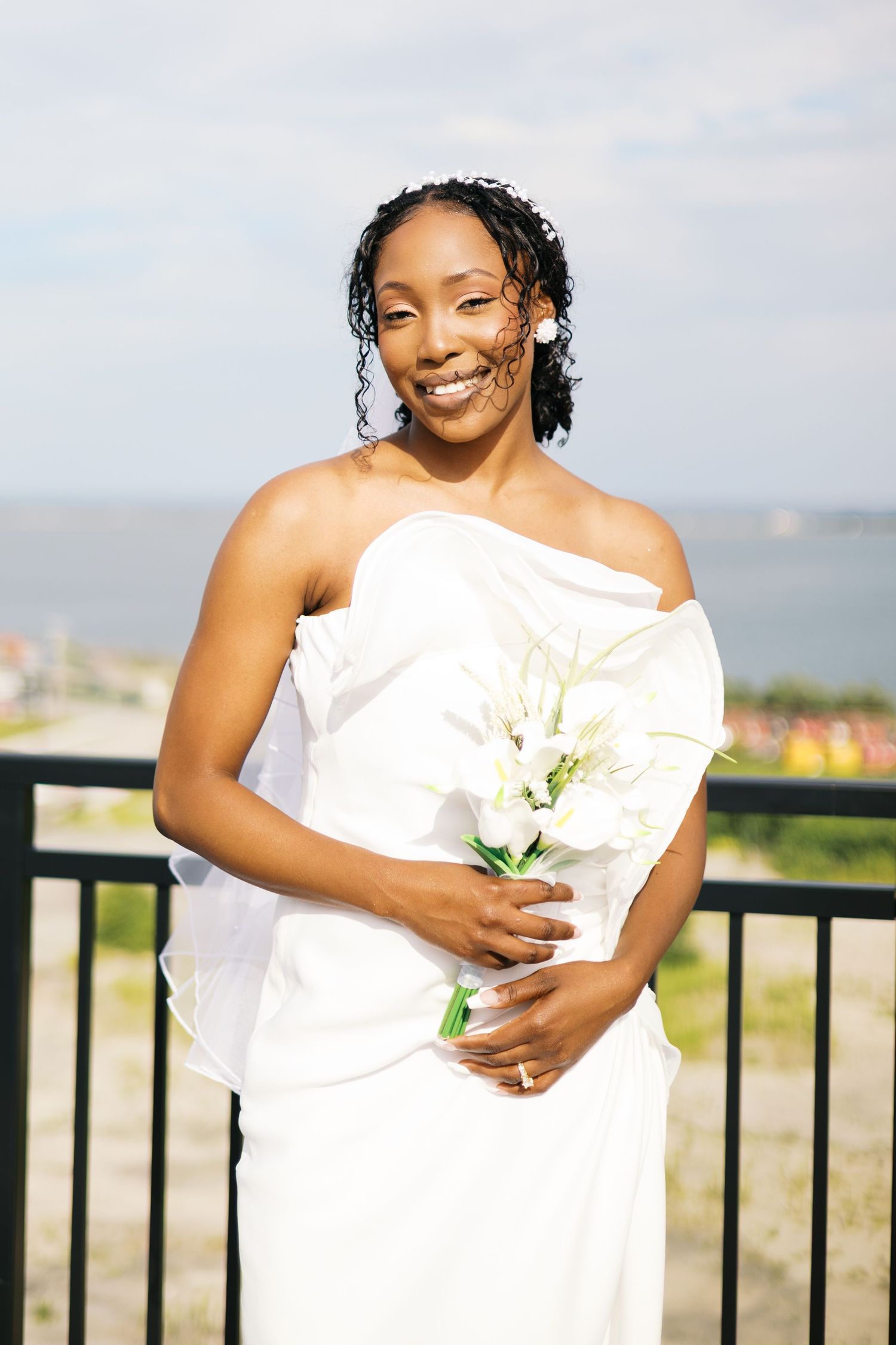 Smiling Black bride in white strapless gown holding white calla lily bouquet outdoors near waterfront.